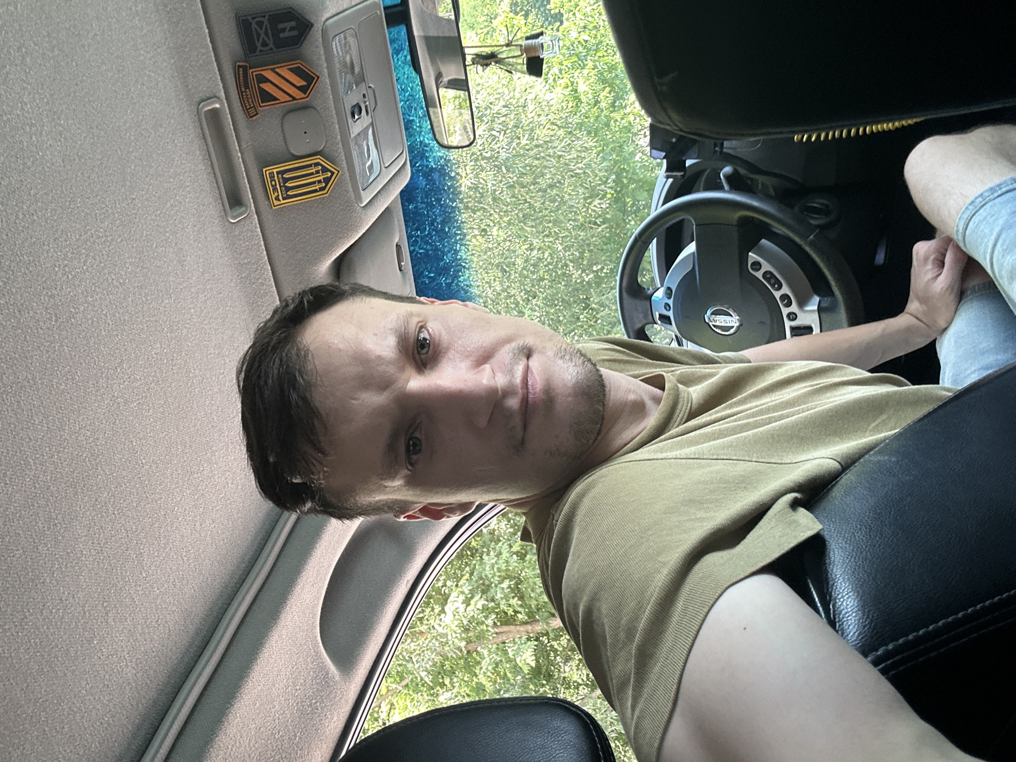 A man is sitting in the driver's seat of a car, looking directly at the camera. He is wearing a green t-shirt and blue jeans. The car's interior is visible, including the steering wheel with a Nissan logo, and the dashboard. There are several patches or emblems attached to the car's ceiling, including one with yellow and blue colors and a trident symbol, possibly related to Ukraine. Outside the car, trees and foliage are visible through the windows. The overall environment suggests a location in a rural or semi-rural area, potentially Makiv, Ukraine, given the context.