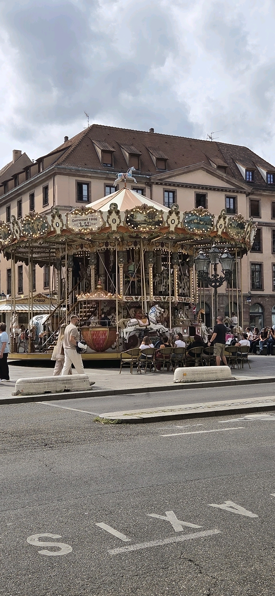 The image depicts a street scene in Strasbourg, France, featuring a carousel as the central element. The carousel, adorned with lights and decorative panels, is set against a backdrop of a multi-story building with a mansard roof and dormer windows. Several people are visible around the carousel; some are seated at tables in front of it, while others are walking nearby. The street in the foreground has markings indicating a taxi stand, with the letters "TAXI" stenciled onto the asphalt. The sky is overcast with clouds.