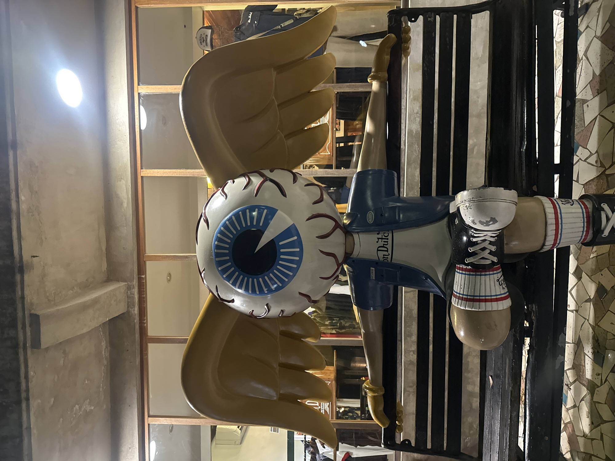 A tall decorative figure is seen indoors in the Ubud District of Indonesia. The figure has a large eyeball with blue and white concentric circles, surrounded by golden wings. The body of the figure is white and blue and has the words "Von Dutch" on its chest. It is wearing a black shoe with white laces. The figure is partially in front of a black metal gate. Behind the figure is a display of various items. The walls are cream-colored. There is a bright light in the upper left of the frame.