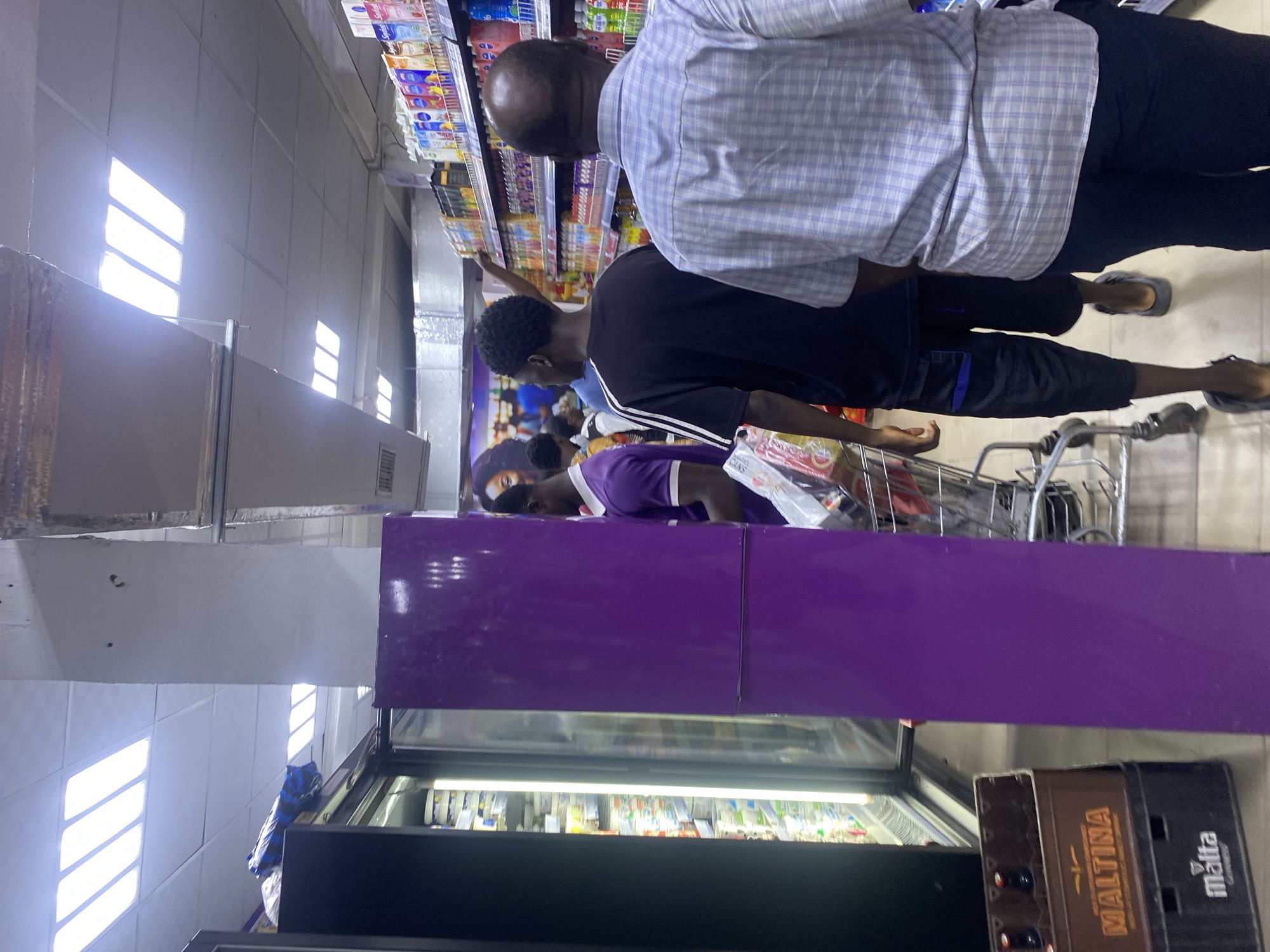 The photograph was taken inside a grocery store, possibly in Lagos, Nigeria. In the foreground are refrigerated display cases with a purple accent panel. Behind the cases are several people in a queue with shopping carts. One man is wearing a light blue checkered shirt, black pants, and sandals. Another man is wearing a black shirt with white stripes on the sleeves and black pants. Further back, another man is wearing a purple striped shirt. Shelves filled with products are visible on the upper right of the frame. The lighting suggests that it is midday or early afternoon.