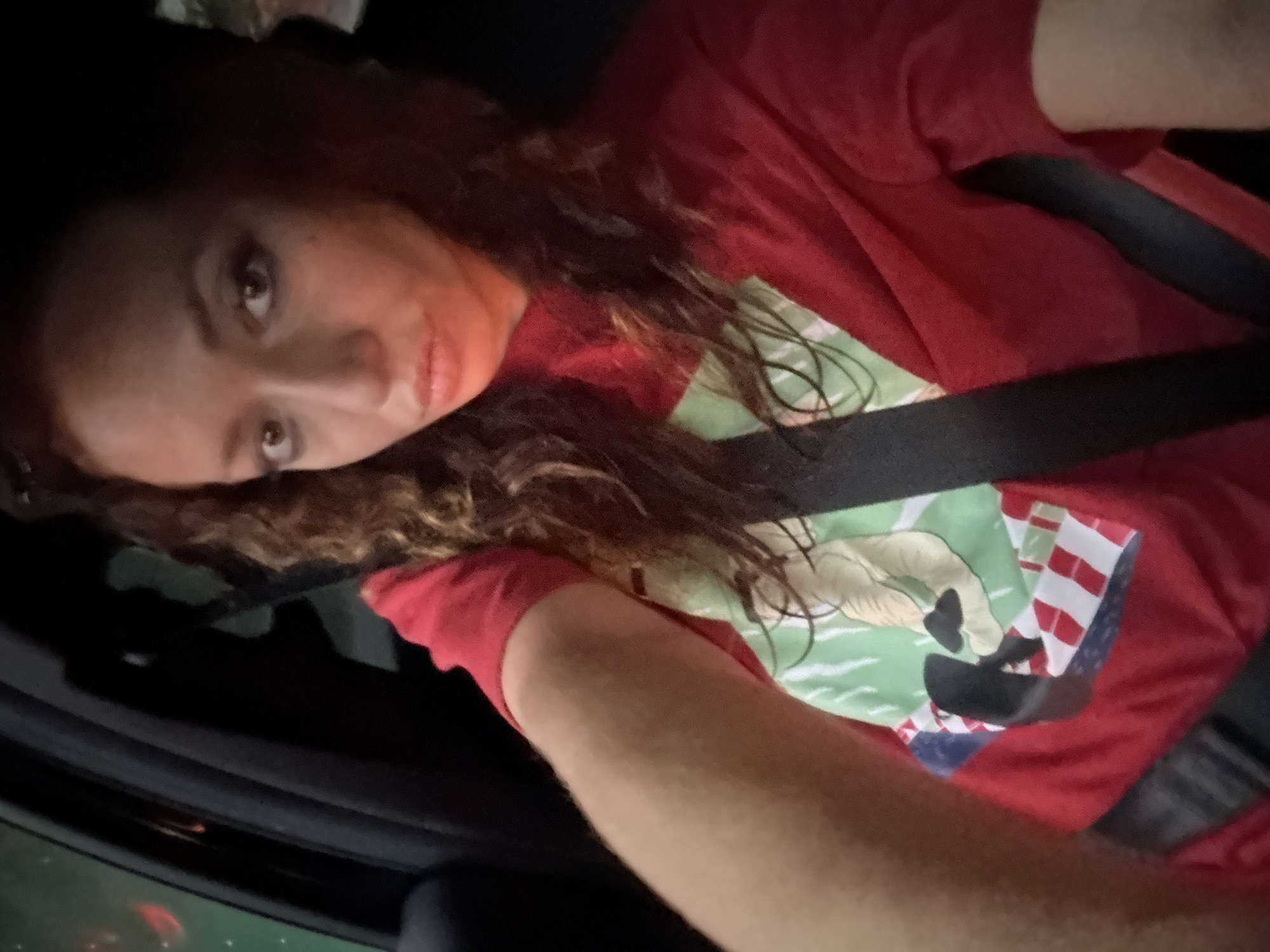 A selfie photograph shows a woman inside a car, wearing a red t-shirt with a graphic of a horse and an American flag. She has brown curly hair and is wearing a seatbelt. The image is slightly tilted, and the lighting is low. The location, Du Già Commune, Vietnam, is not discernible from the visual information in the image.