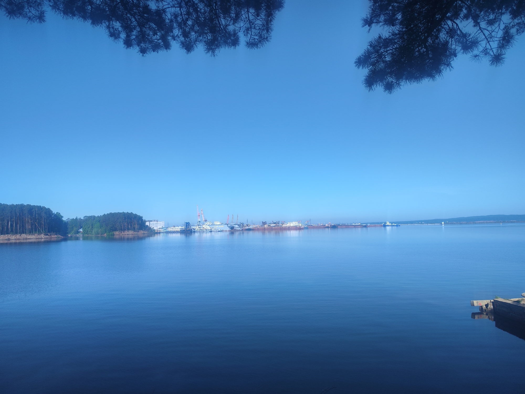 A serene, daytime shot captures a waterfront scene in Unknown, Unknown. The image is split into three distinct horizontal sections. The upper third is a clear, bright blue sky framed by the dark, blurred silhouette of tree branches and foliage at the top. The middle third consists of a calm body of water, reflecting the sky. Along the horizon line, a cityscape or industrial complex is visible, featuring various buildings, cranes, and structures. The lower third of the image is the foreground water, which darkens to a deep blue. A concrete structure juts out into the water on the right side of the image. The overall impression is one of stillness and tranquility.