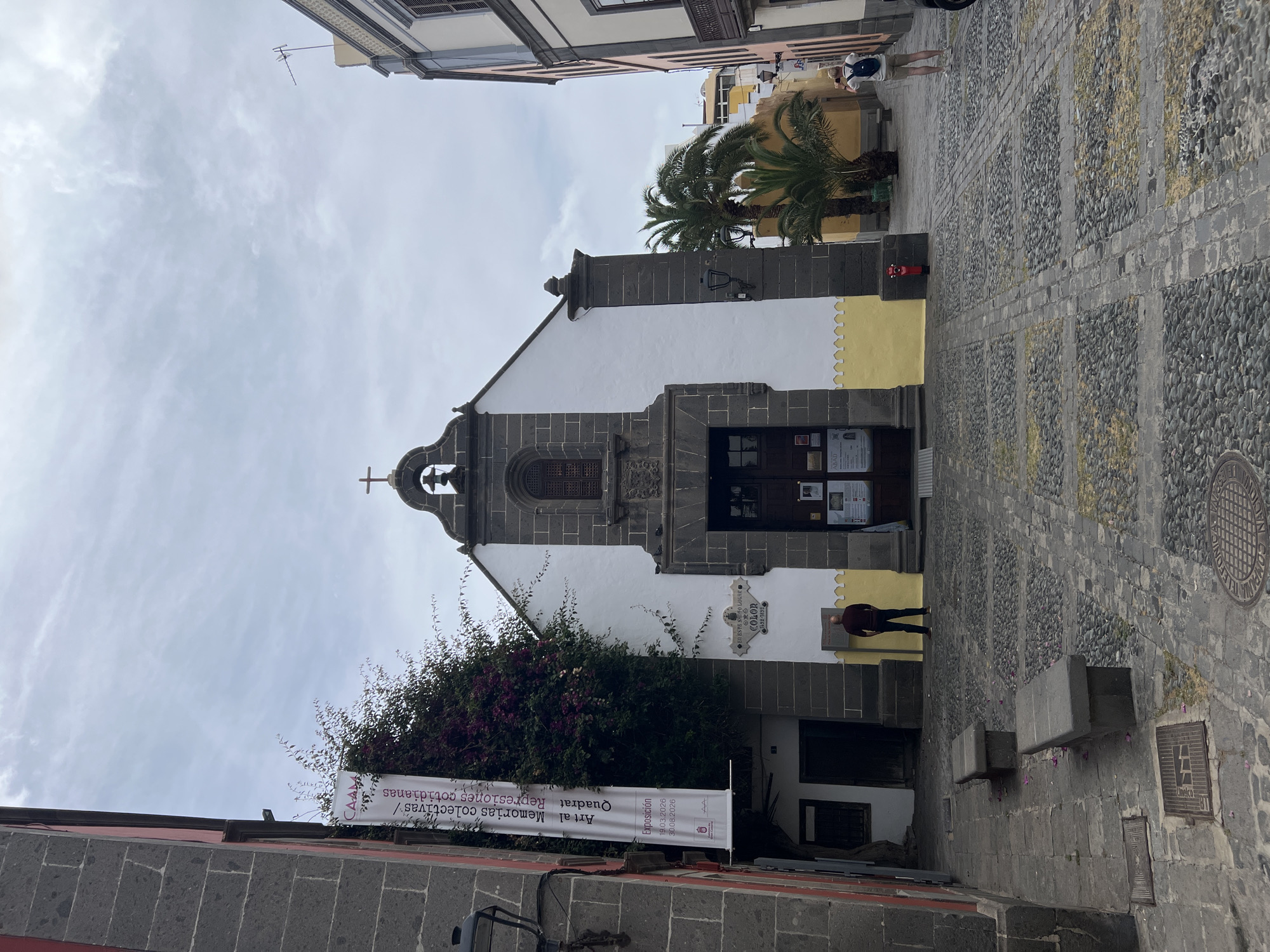 Here is a detailed description of the image:
The image depicts a street scene in the city of Las Palmas De Gran Canaria, Spain, featuring a church situated between buildings. The architecture of the church is characterized by a white facade with dark stone detailing. It has a bell tower surmounted by a cross. A banner hangs on the left side of the church, partly obscured by foliage, with text reading "Art al Memorias colectivas" and exhibition dates "19.03.2026 - 30.08.2026." A small plaque with the word "COLOR" is mounted on the church wall.
The street is paved with patterned stones. A red fire hydrant is visible on the right side of the church. A few people are present: one is walking near the church entrance, and others are further up the street. The buildings on either side of the church show a mix of architectural styles and colors. Palm trees are also visible.
The sky appears overcast. The scene is typical of an urban environment with a mix of historical architecture and modern elements.