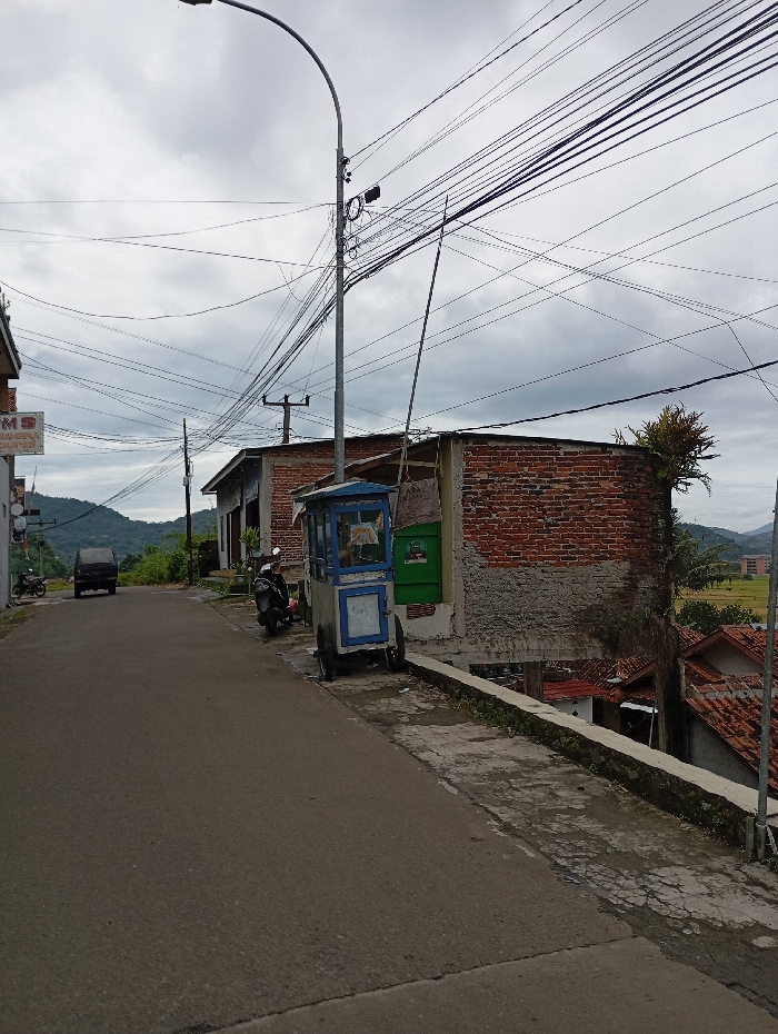 The image shows a street in Kecamatan Kutawaringin, Indonesia, at the coordinates -6.987 latitude, 107.531 longitude. The scene features a paved road with a small food cart parked on the right-hand side. The cart is painted blue and has a white sign with text that is not visible. A motorcycle is parked in front of the cart. Behind the cart is a brick wall. The wall is made of red bricks and painted white. Behind the wall, there is a small green roof and a patch of green grass. Above the street is a cloudy sky with a streetlight extending from the left side of the frame and a large number of electrical wires that crisscross the scene. The image was captured from the point of view of a person standing on the side of the road looking down the street. There are no people in the image. The street is mostly empty, with only a single dark-colored car visible in the distance.