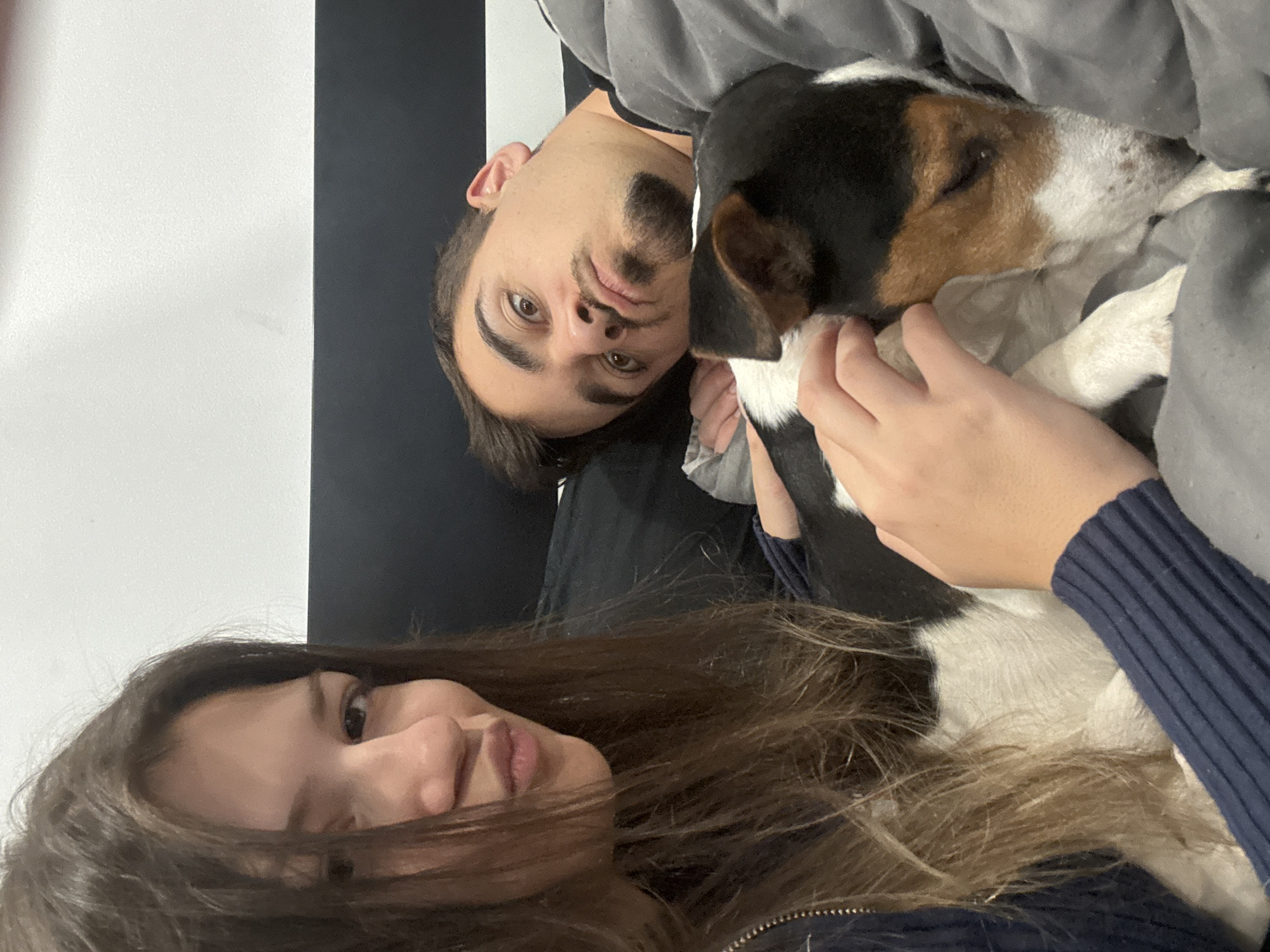 The image shows two people and a dog. A young woman with long brown hair is in the foreground, her face visible. Behind her, a man with dark hair and a beard is partially visible. He is holding a tricolor dog (black, white, and brown) wrapped in a grey blanket or fabric. The dog appears to be asleep or resting comfortably. The background consists of a wall with alternating black and white stripes. The location in Sofia, Bulgaria, cannot be determined from the image alone.