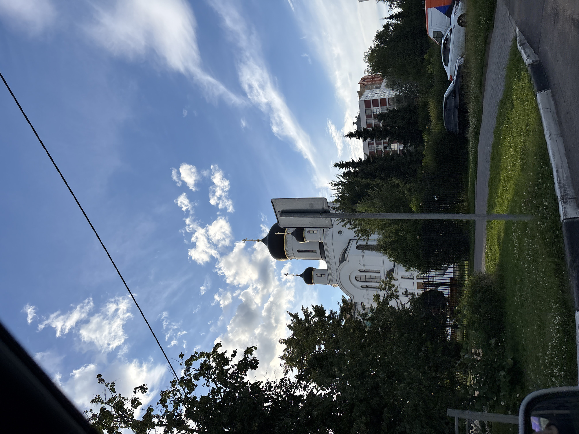 The image captures a clear day in Mytishchi, Russia, featuring a white Orthodox church with black domes, partially obscured by a large tree on the left. In the background, several modern apartment buildings are visible amongst dense green trees. The scene includes a black car parked on the side of the road, a white car, and a white and orange van. A black wire stretches diagonally across the bright blue sky, which is adorned with scattered white clouds. The foreground shows a grassy area with small white flowers and a paved walkway.