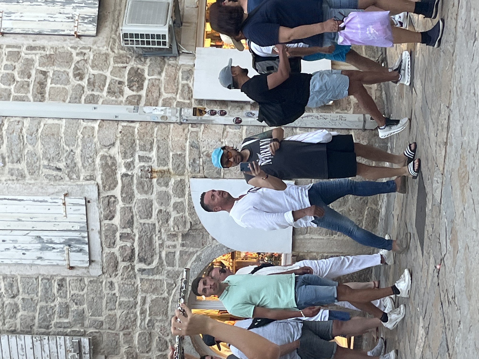 The image captures a group of men walking on a cobbled street in Budva, Montenegro. The architecture suggests an old town setting, with a stone building featuring wooden shutters and an outdoor air conditioning unit visible. One man in the foreground is playing a flute, while others are engaged in conversation or looking at their phones. The attire of the individuals indicates a casual, summer atmosphere. The scene appears to be a candid street photograph, with the focus on the people and their activities as they navigate the urban environment.
