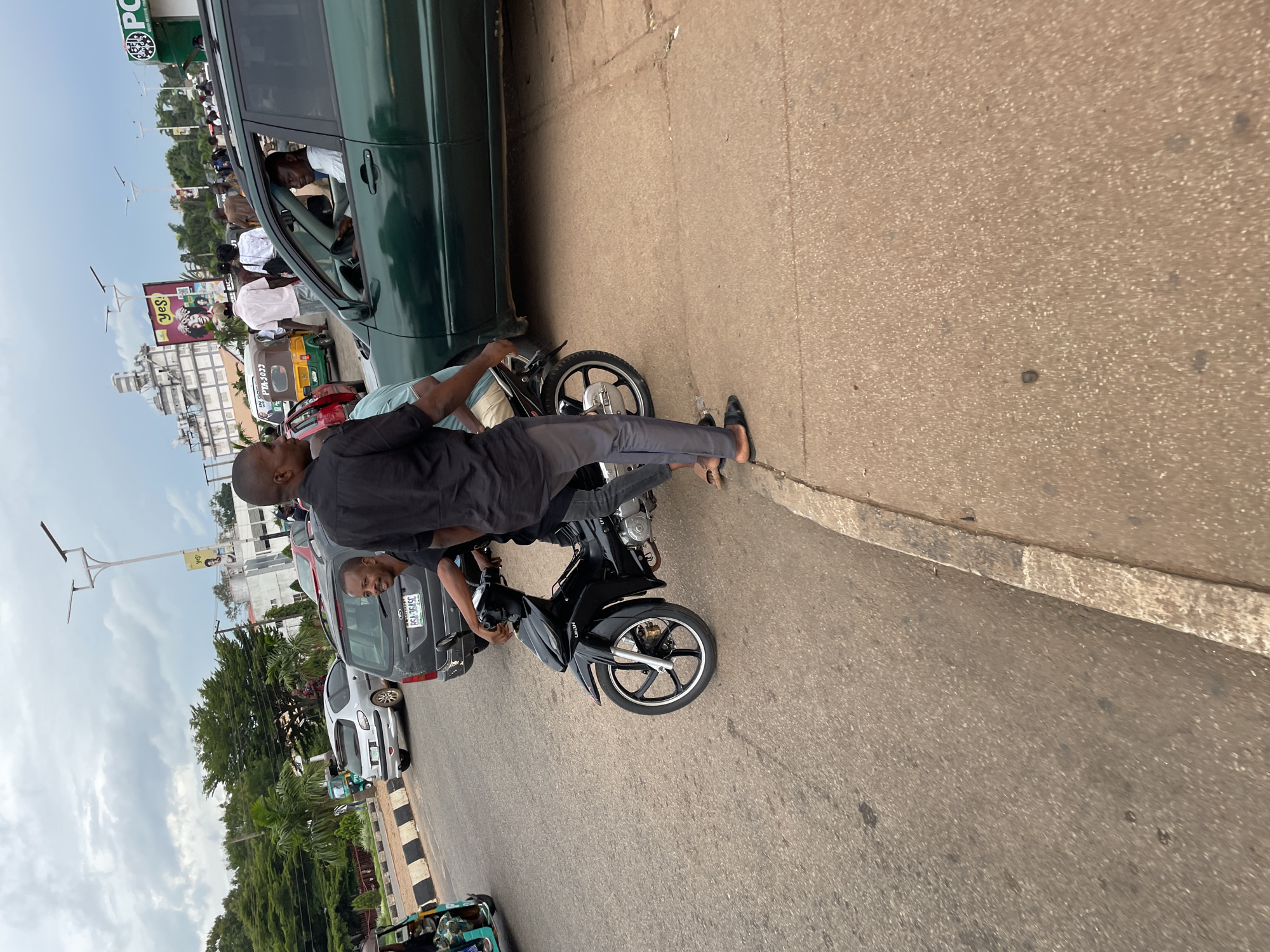 In the city of Jos, Nigeria, a black motorcycle is parked on the side of a road with two men on it. The man in the back is wearing a black shirt and gray pants, while the man in the front is wearing a light blue shirt. A green SUV is parked next to the motorcycle. Other vehicles and pedestrians are visible in the background, along with buildings and billboards. The sky is cloudy.