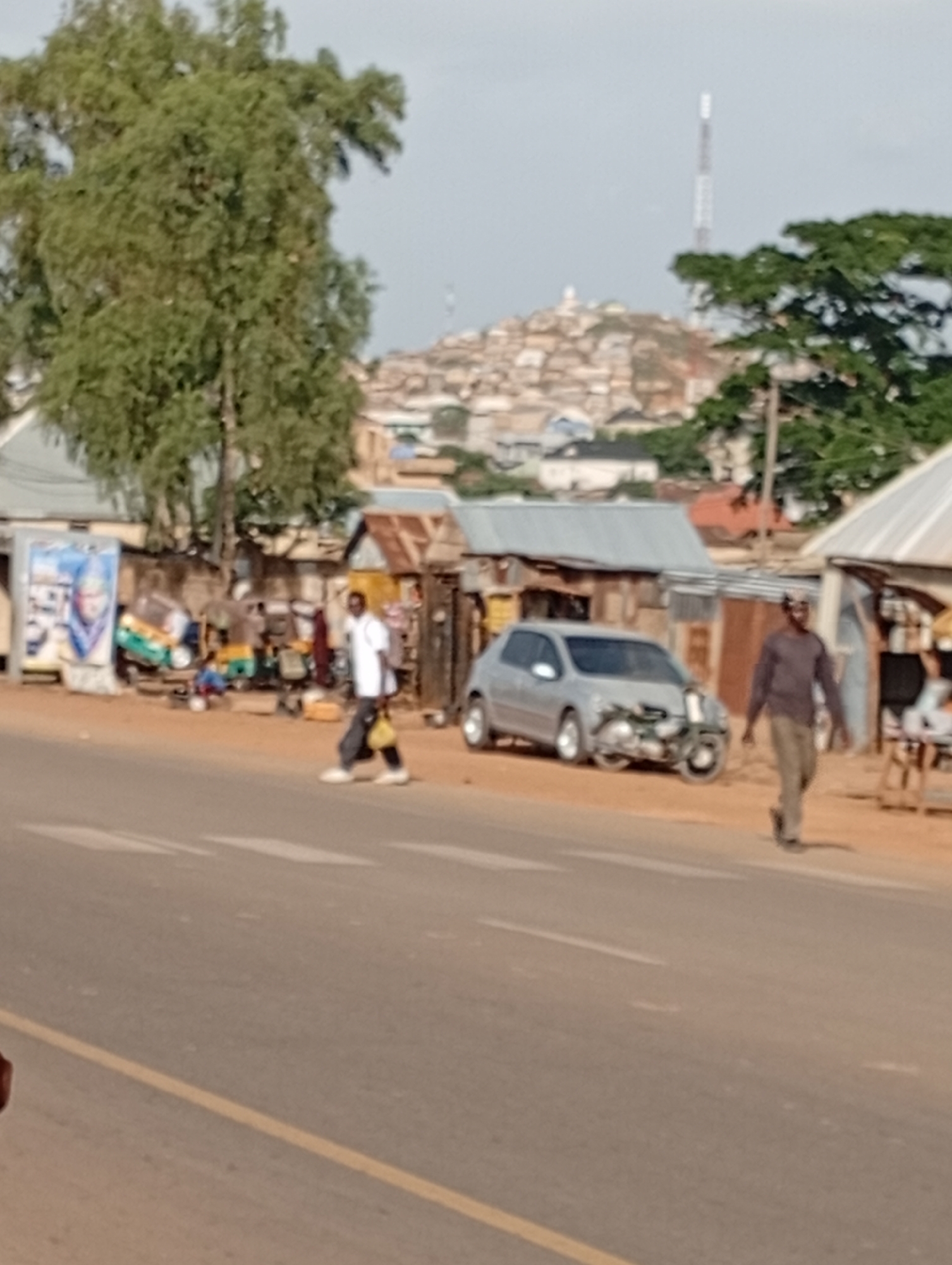 A medium shot captures a street scene in Rafin Par, Nigeria, under a hazy sky. In the foreground, a paved road with a yellow line is visible. Two men are walking on the side of the road. The man on the left is wearing a white shirt and dark pants, carrying a yellow bag in his right hand. The man on the right is wearing a dark long-sleeved shirt and light-colored pants.
Behind them, a silver car, possibly a hatchback, is parked on the side of the road. Its front end appears to be damaged. The background reveals a row of small buildings with corrugated metal roofs, some appearing to be market stalls or shops. A large tree with green foliage stands to the left of the buildings. Further in the distance, a densely populated hillside is covered with numerous buildings, and a tall transmission tower is visible against the sky. The overall impression is of a busy urban or semi-urban environment.