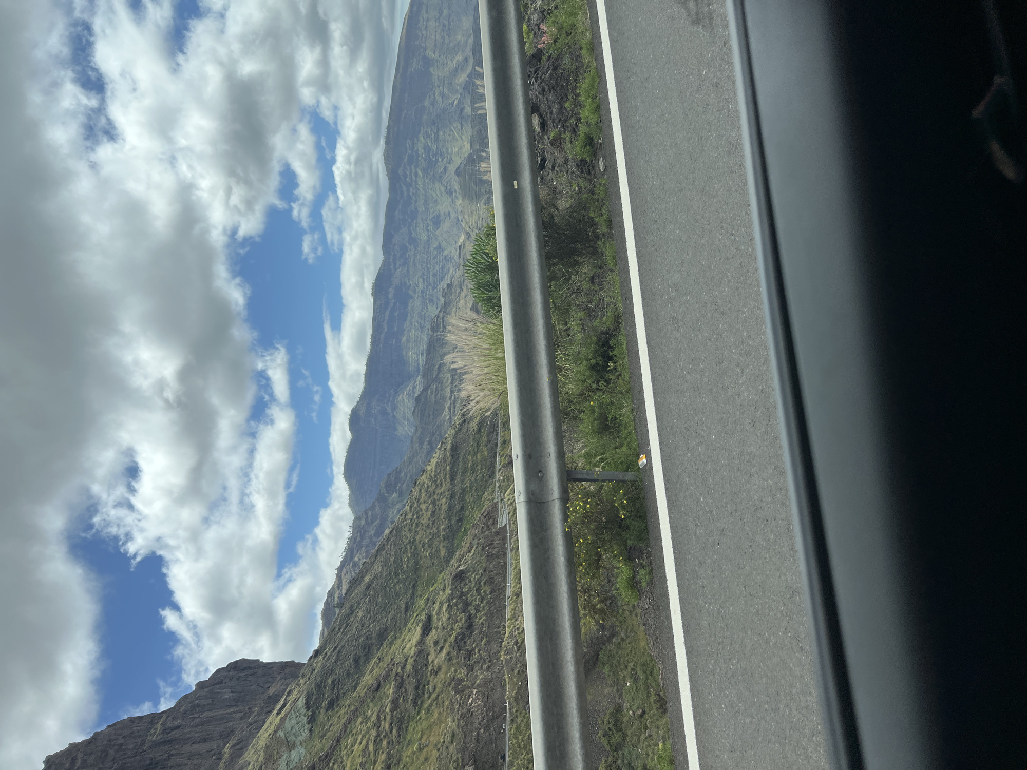 The photograph, taken from inside a vehicle, shows a roadside scene in Mogán, Spain. A grey guardrail separates the road from a steep, green, mountainous terrain. The road is asphalt, marked with a white line along the edge. In the distance, layered mountain ridges are visible, ascending into a sky partially obscured by white, puffy clouds with patches of blue sky showing through. The view suggests a high altitude or elevated road position. The vehicle frame is visible, partially blocking the right portion of the photograph. The composition emphasizes the contrast between the man-made road and the natural, rugged landscape.