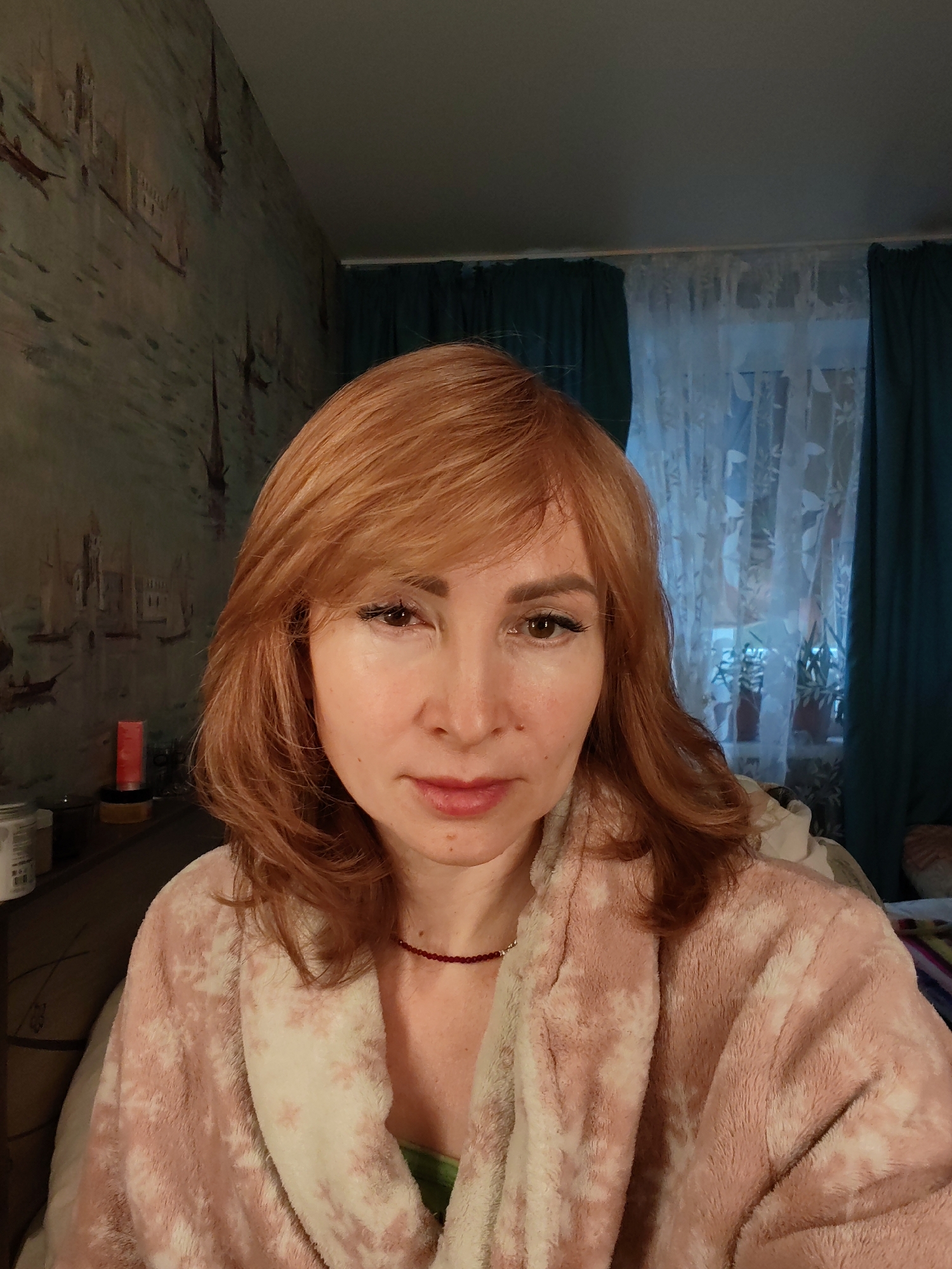 The image depicts an adult female positioned centrally in an indoor environment within Chaikovsky, Russia. She has reddish-brown, shoulder-length hair with a fringe, fair skin, dark brown eyes with noticeable eyelashes, and closed, reddish-pink lips, looking directly forward. She is wearing a soft, fluffy robe with a pink and white pattern. A green garment and a thin, dark red beaded necklace are visible below the robe at her neckline. The background to her left features wallpaper with a faded illustration of a European city scene, including buildings and boats on water. Below this wallpaper, on a horizontal surface, are several objects: a tall, cylindrical red item, a dark circular container, and a white jar with indistinct markings. To her right, a window is visible, covered by white sheer curtains with a patterned design, flanked by darker teal curtains. Faint outlines of potted plants are discernible on the windowsill behind the sheer curtains. The lighting in the scene is moderate, with the window area appearing as a source of ambient light.