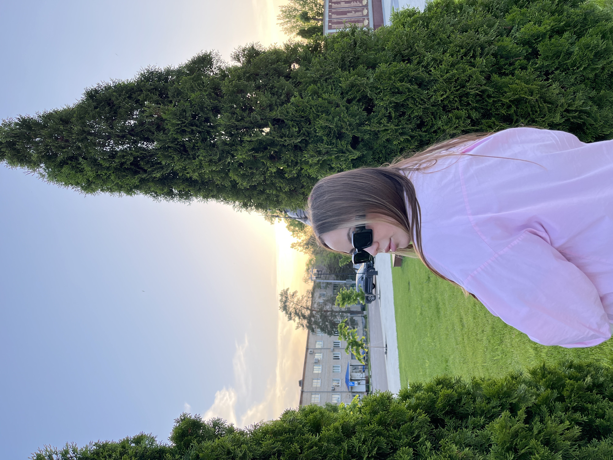 A woman wearing sunglasses and a pink jacket is standing in a park in Novopokrovskaya, Russia. She is positioned between two bushes, with a tall, thin tree visible in the background. Behind the tree, there is a building, a sidewalk, grass, cars, and an illuminated sky.