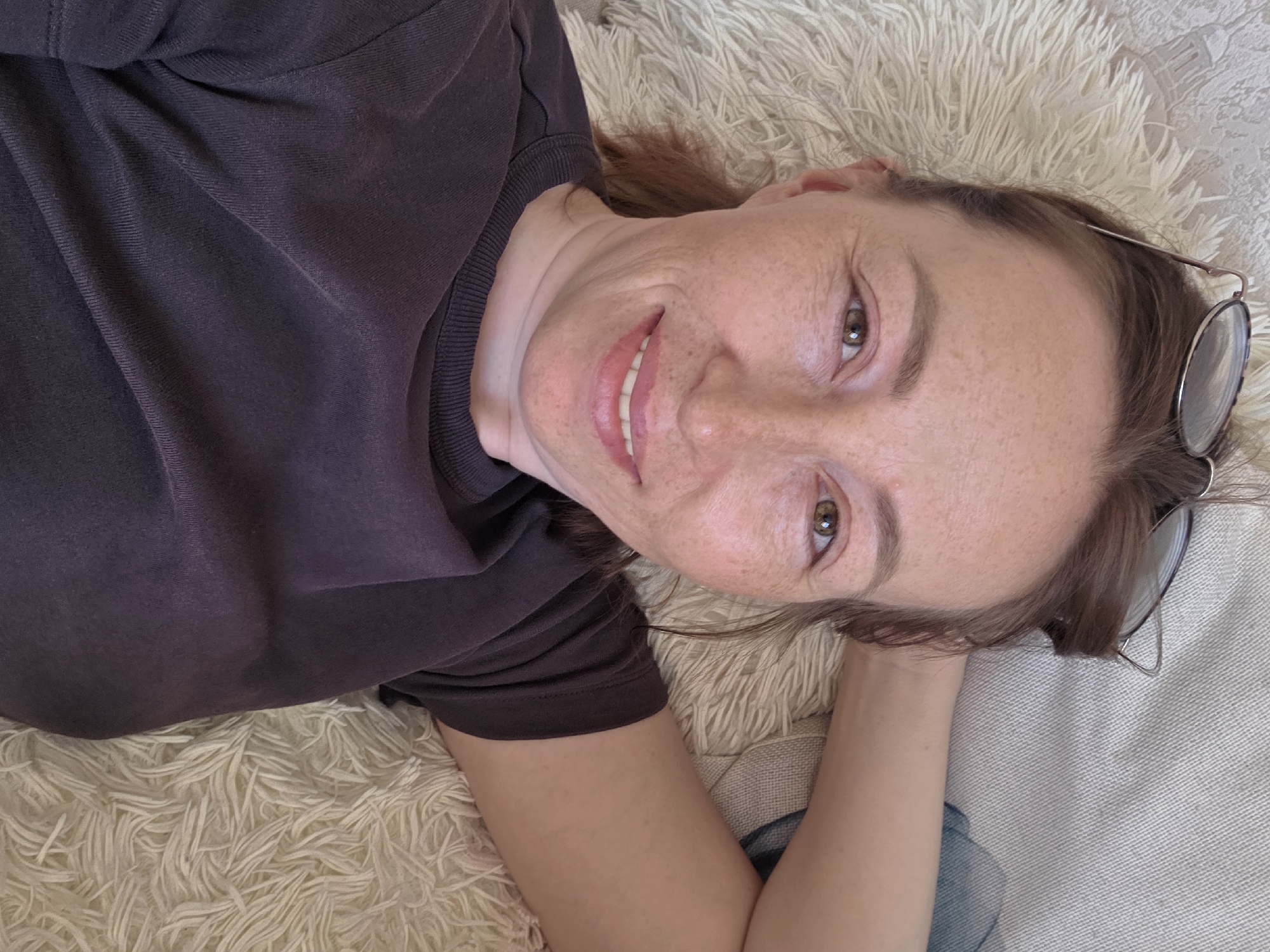 A fair-skinned woman with red hair and freckles lies on her side on a white, fluffy rug or blanket. She wears a dark purple t-shirt and smiles at the camera. A pair of glasses rests on the rug near her head. Part of a light-colored couch and what appears to be a blue article of clothing are visible in the lower right corner. The location is presumed to be an interior setting, potentially in the city of Chaikovsky, Russia.