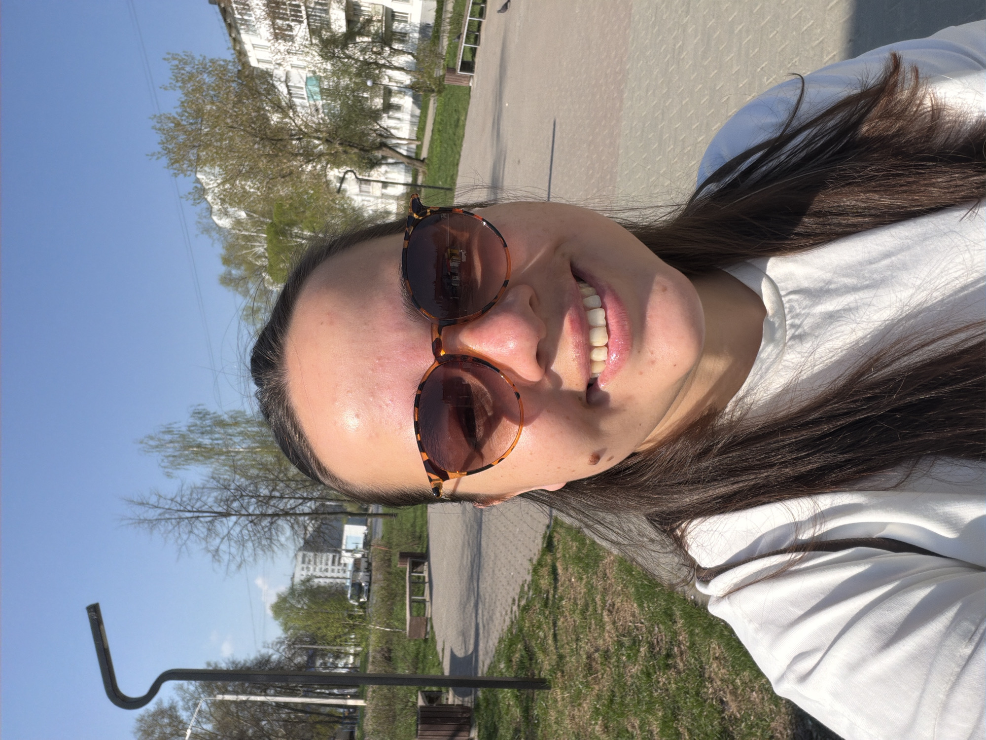 A young woman poses for a selfie outdoors in Chaikovsky, Russia. The photo is taken at an angle, showing her face, part of her torso, and the background environment. She wears sunglasses with brown tortoiseshell frames and a white top. Her hair is long and dark brown. She smiles, revealing her teeth. Behind her is a park with trees, grass, a paved walkway, benches, and a building in the distance. A streetlight or other pole is visible on the left side of the frame. The sky is blue with few clouds.