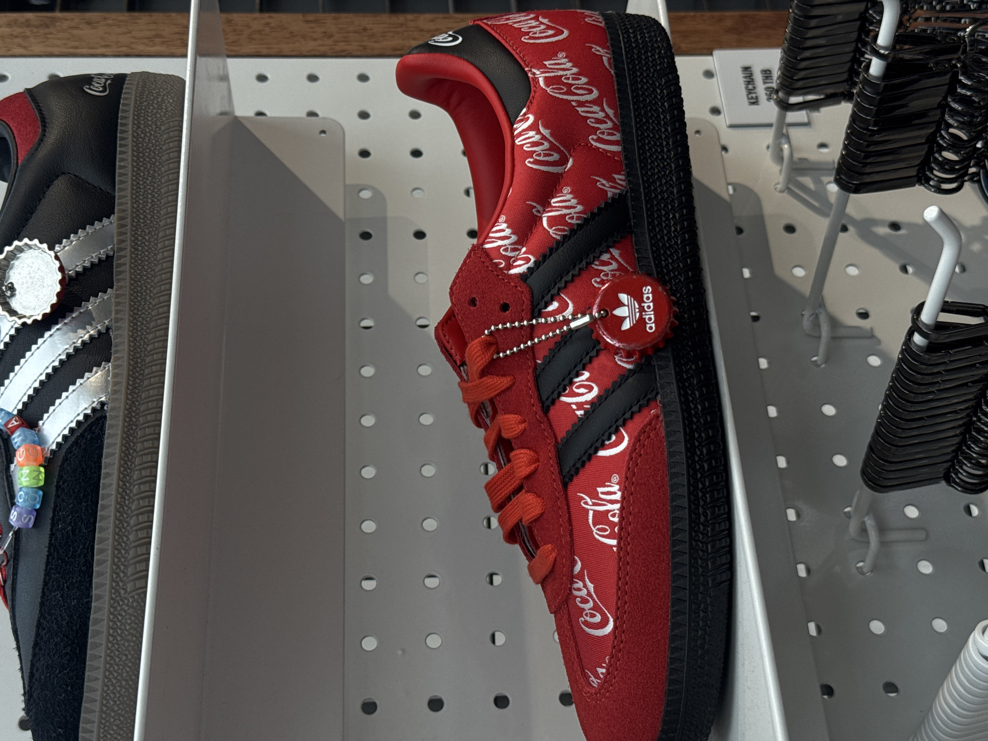 The image shows two Adidas sneakers, one black and one red, displayed on a white shelf against a pegboard background. The red sneaker, in the foreground, is a collaboration with Coca-Cola, featuring a repeating Coca-Cola logo pattern. It has black Adidas stripes and a black sole. A red keychain with the Adidas logo and a Coca-Cola bottle cap design hangs from the laces. The black sneaker, in the background, also appears to be a Coca-Cola collaboration, with a silver Adidas logo and a red heel tab. Colorful beaded charms are attached to its laces. The scene is indoors, likely in a retail store, given the display shelving and the presence of a price tag for a keychain that reads "KEYCHAIN 250 THB". The background suggests a modern retail environment, common in shopping centers. The scene is devoid of any people.