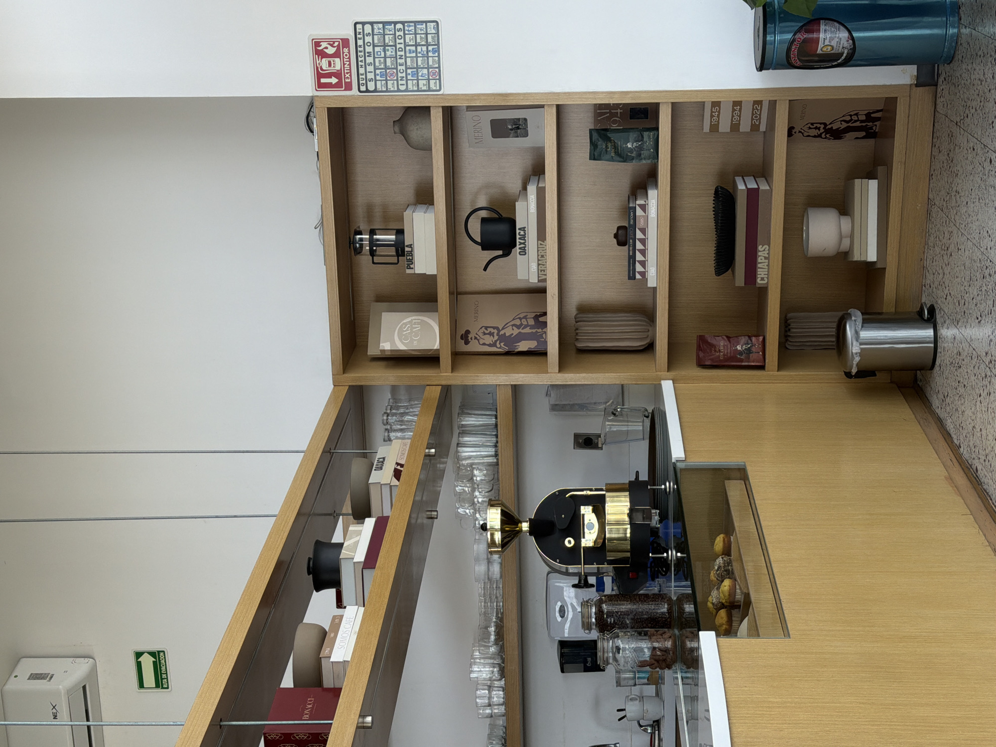 This media file captures the interior of a well-organized coffee shop or a coffee-themed retail space, featuring a prominent wooden shelving unit and a counter. The shelving unit is filled with various coffee-related items, books, and decorative objects.
On the top shelf, a gray vase, a book titled "MERINO," and a green bag labeled "CAF 1943" are visible. The middle shelf displays a French press next to books, including one labeled "PUEBLA," a black kettle, and books titled "OAXACA" and "VERACRUZ." Other books on this shelf include "BONACCIA," "LAROUSSE CAFE," and "CIAO." The bottom shelf features a book titled "CASA DEL CAF," another "MERINO" volume with an illustration, a reddish-brown bag also labeled "MERINO," and a stack of books marked with the years "1945, 1994, 2022." Books labeled "CHIAPAS" are also present.
Below the main shelving, a counter holds a distinctive golden and black coffee machine, several glass jars filled with coffee beans or grounds, and a display case containing small pastries or muffins. Numerous clear glasses are neatly arranged on the counter.
On the white wall, several emergency signs are visible: a red "EXTINTOR" (Fire extinguisher) sign pointing downwards, a blue and white sign titled "QUE HACER EN: SISMOS INCENDIOS" (What to do in: Earthquakes Fires) with pictograms detailing emergency procedures, and a green "RUTA DE EVACUACION" (Evacuation Route) sign with an upward arrow. A white box labeled "NEX" is also mounted on the wall. The floor is tiled. A silver trash can is located on the right side of the image. The Spanish text on the signs and the names of Mexican states on the books (Puebla, Oaxaca, Veracruz, Chiapas) suggest a location in Mexico, aligning with the provided context of San Bernardino Tlaxcalancingo. No individuals are present in the scene.