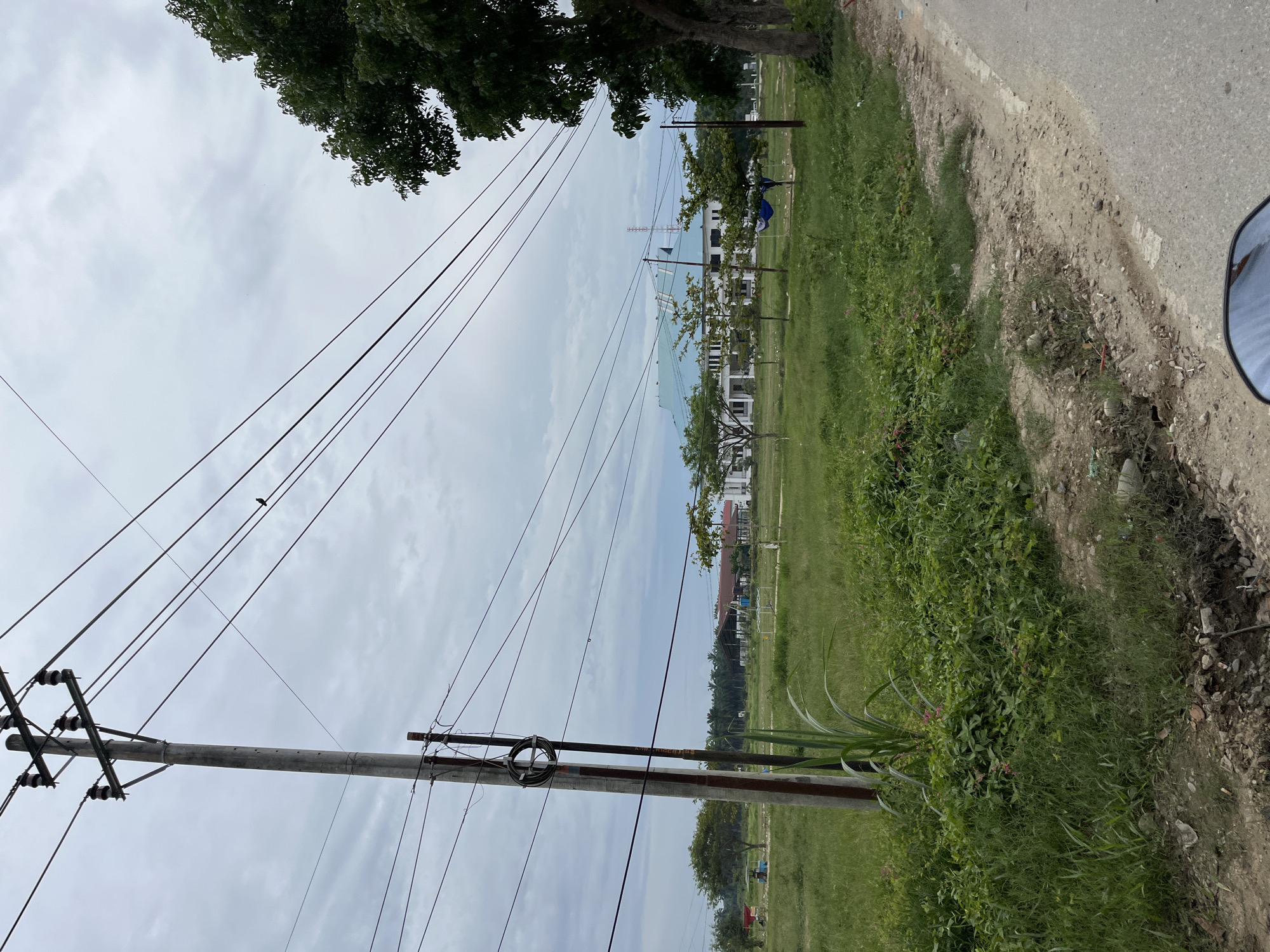 The image depicts an outdoor scene in Bundar, Indonesia, with a focus on a utility pole and its overhead wires. The pole, made of concrete, stands to the left of the frame and supports multiple electrical wires. A coil of black cable is attached to the pole. In the background, a white building with a teal-colored roof is visible, surrounded by green grass and scattered trees. The sky is overcast with clouds. A paved road runs along the right side of the image, and the edge of what appears to be a motorcycle's rearview mirror is visible in the lower right corner. The ground in front of the building is grassy, with some patches of dirt and debris.