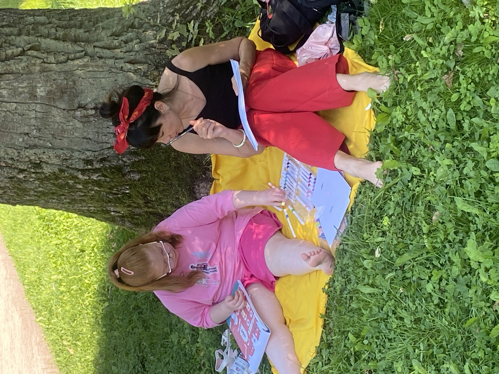 Two individuals are seated on a yellow blanket in a grassy area, likely near a large tree. The person on the left, with ginger hair pulled into a ponytail, wears a pink t-shirt and pink shorts, and glasses. They are holding an open book or coloring book and appear to be coloring or drawing with markers. To their right, a person with dark hair tied up and a red bandana on their head, wears a black tank top and red pants. They are holding a pen and a piece of paper, also appearing to be engaged in drawing or writing. A collection of markers is laid out between them. A black backpack and a pink cloth are visible to the right of the person in red. The ground is covered in lush green grass and clover. The image was taken in the city of Pushkin, Russia.