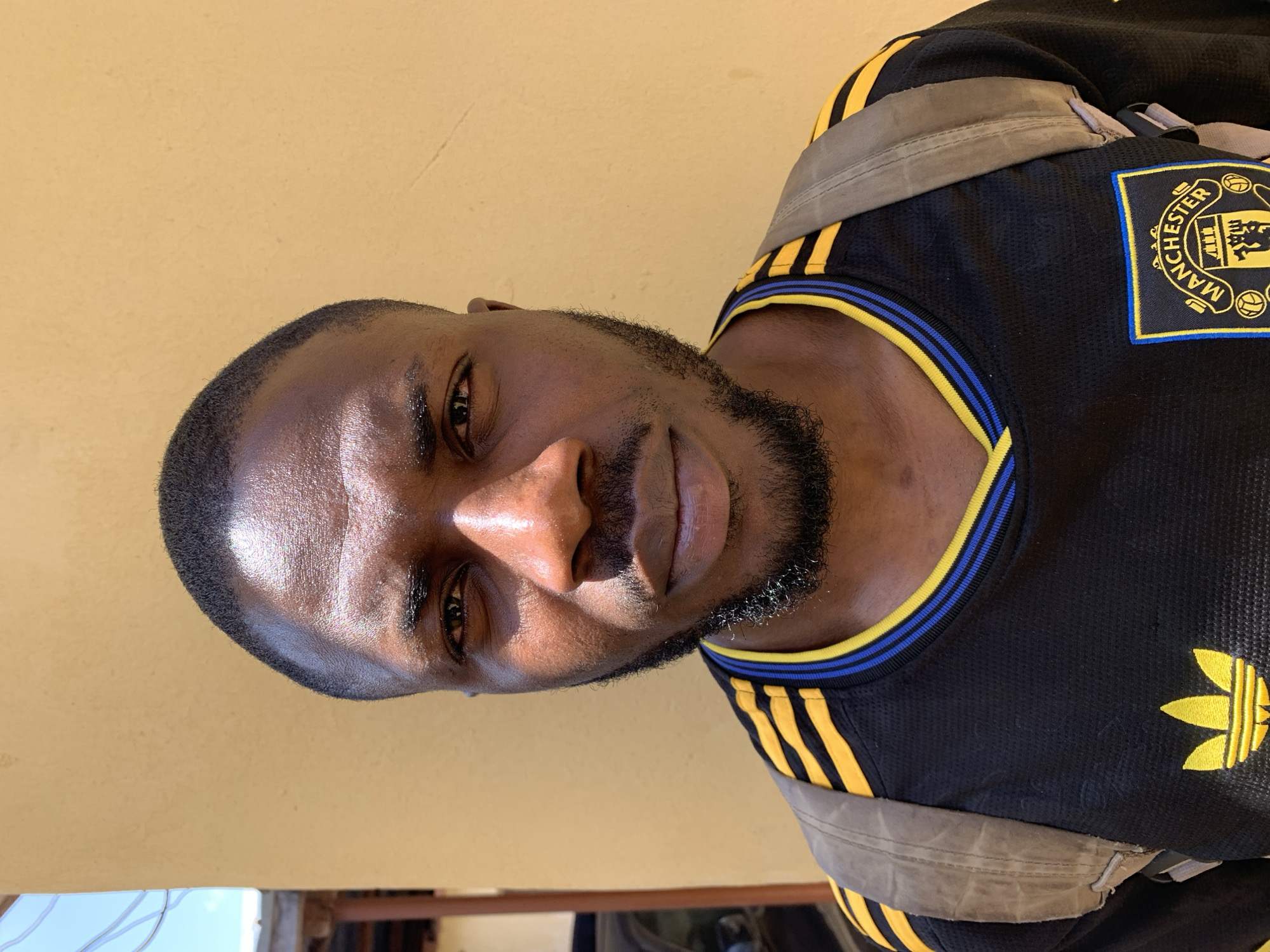 A man with dark skin and a beard is wearing a black Manchester United Adidas jersey with yellow stripes and collar detail and a backpack, standing against a pale yellow wall. The man is in the city of Rafin Par, Nigeria.
