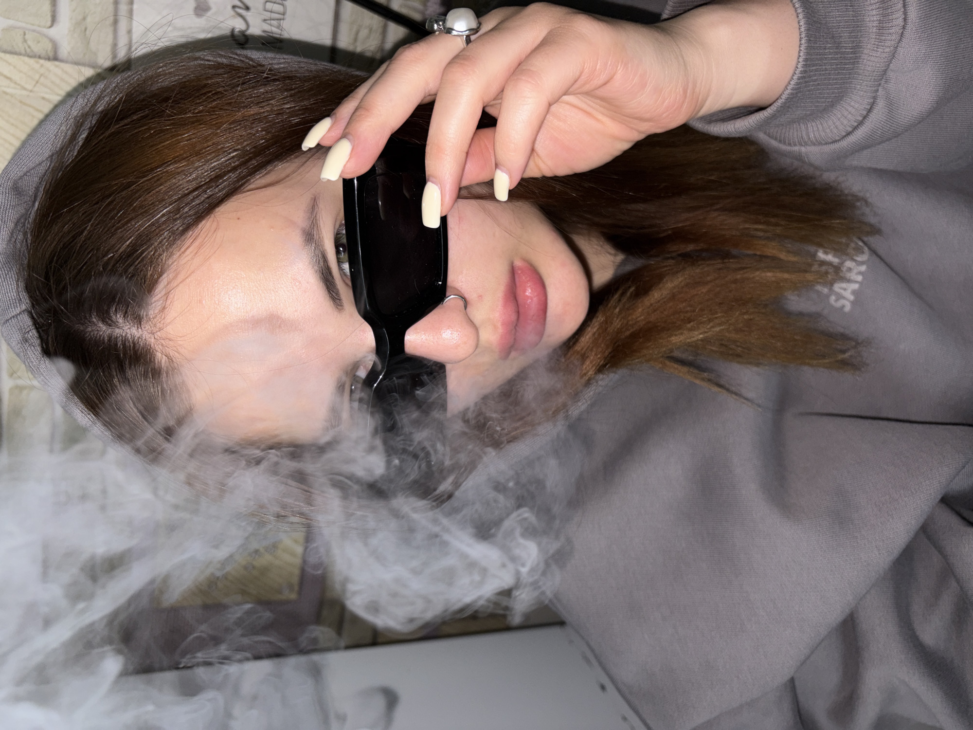 The image depicts a person wearing a grey hooded sweatshirt and sunglasses. They have brown hair and a silver ring on their finger. There is white smoke obscuring the lower portion of the image. A brick-patterned wall is visible in the background. The location is not discernible as Port-Katon, Russia, based on the provided image.