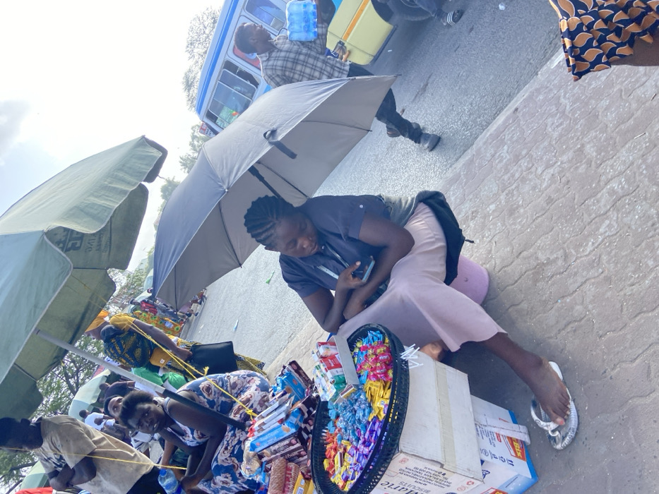 This image, rotated clockwise, depicts a bustling street market scene in Dar es-Salaam, Tanzania, under bright daylight conditions. In the foreground, a young woman is seated on a pink cushion, sheltered by a dark grey umbrella. She wears a dark short-sleeved shirt and a light pink skirt, and is focused on a smartphone held in her hands. In front of her, a black oval tray overflows with an assortment of colorful candies and chocolates, alongside several cardboard boxes containing packaged goods. The brand name "MARIE" is visible on one of the boxes.
Further back, several other female vendors are visible, also sitting on the ground with their merchandise, mostly under large light-green umbrellas providing shade. One of these umbrellas faintly shows the text "TVC." Another vendor in a blue and white patterned dress is seen looking towards the camera. In the background, a man in a plaid shirt stands near a blue and yellow bus, likely a daladala, holding a large blue plastic water bottle. The ground is paved, indicating an urban roadside setting. The prevalence of umbrellas suggests a sunny day and the need for protection from the heat. The scene captures the active commerce of a typical roadside market.