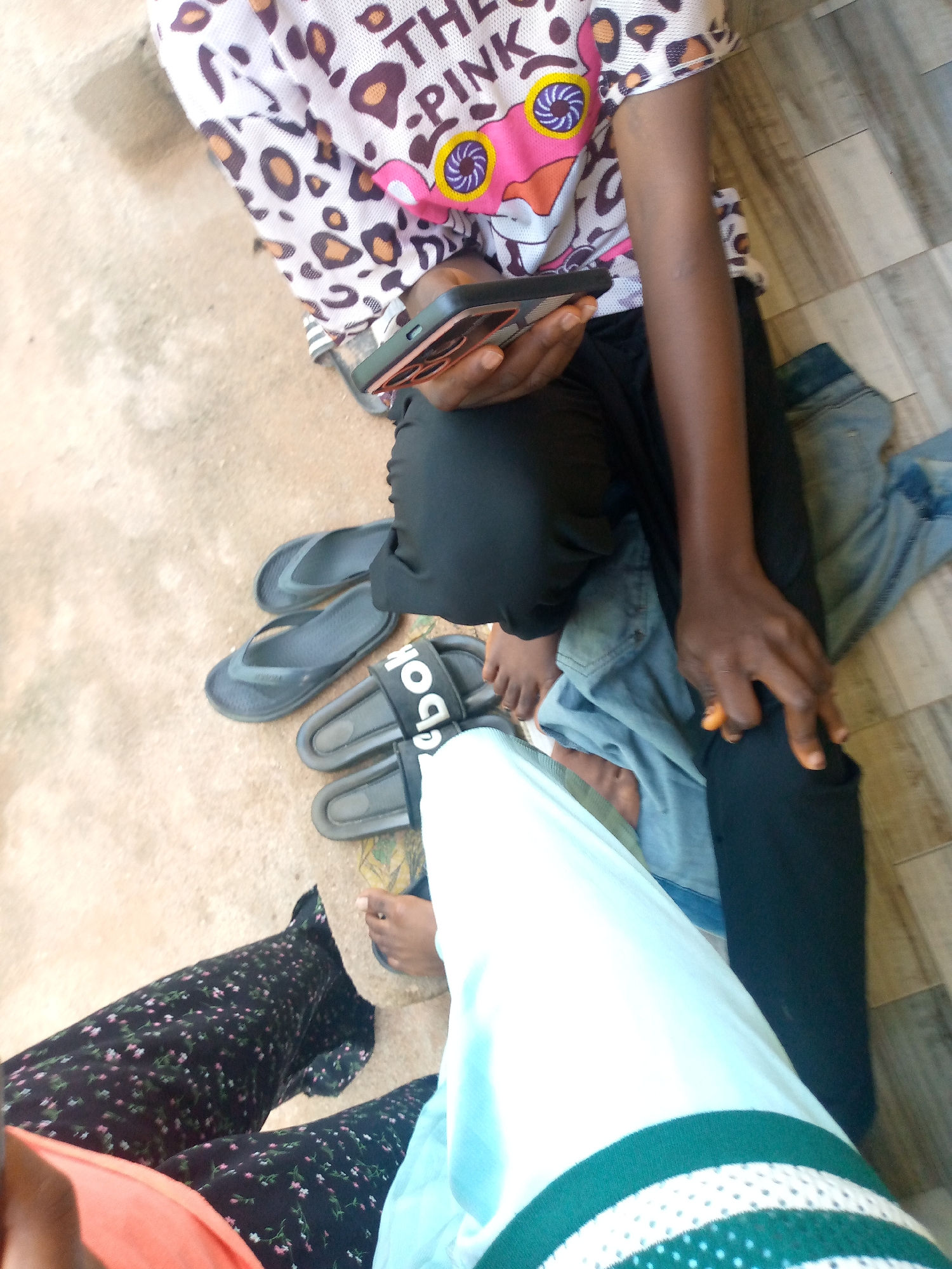 In Jauro Yinu, Nigeria, two individuals are seated on a concrete and wooden plank surface. One person, wearing a white t-shirt with a leopard print pattern and text that includes "THE PINK," is holding a smartphone. This individual is also wearing black pants and has a pair of black flip-flops nearby. The other person is wearing a black floral print skirt and an orange top. Their legs are crossed, and a light blue garment with green and white stripes is visible. Several other pairs of flip-flops are scattered on the ground. The lighting suggests it is daytime.