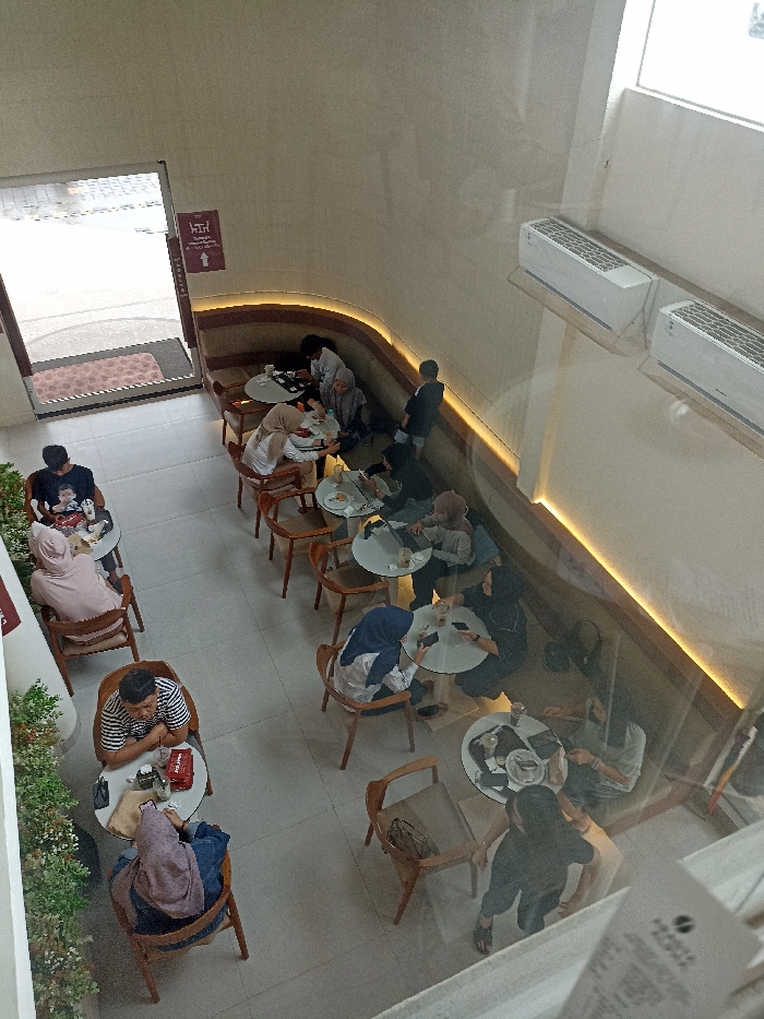 The image shows a cafe located at the coordinates -7.42 latitude and 109.245 longitude in Purwokerto, Indonesia. The cafe has a curved, wooden banquette along the wall, and a large window overlooking the street. The cafe has white tiled floors, white walls, and is lit by recessed lighting. There are 10 people seated at tables, and 2 people are standing. The people are sitting on chairs and eating or drinking, and some are engaged in conversation. The tables are round, and the chairs are wooden. On the tables are plates, cups, and glasses. The cafe is modern and stylish. There are two air conditioning units on the wall. There are also several plants growing in the cafe. The cafe is empty, but the lights are on, suggesting that the cafe is open. The cafe is very clean. The image was taken from above, and there are no shadows. The view from above gives a sense of the overall layout of the cafe. The cafe appears to be popular and well-used.