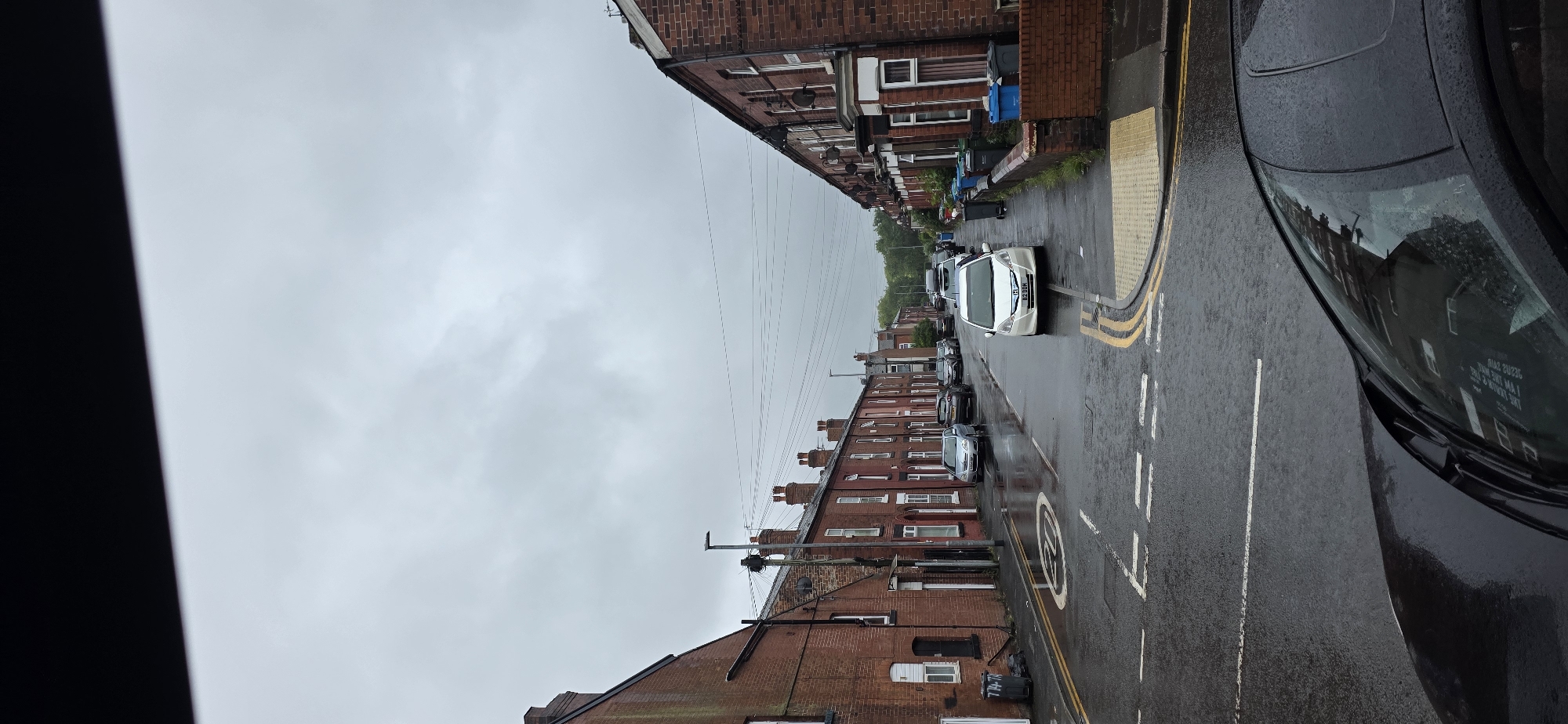 This is a vertical, full-shot photograph of a residential street in Manchester, United Kingdom, on an overcast day. The street is lined with terraced houses made of red brick, characteristic of Northern England. The road is wet, suggesting it has recently rained or is still raining.
On the left side of the street, a red brick terraced house with white-framed windows is visible. Two grey refuse bins are placed on the pavement in front of it. The street has a speed limit sign on it, indicating 20 mph, and double yellow lines marking parking restrictions.
Parked cars line the right side of the street, including a white hatchback with license plate "BZ DOH", a silver sedan, and a darker colored SUV. Several other cars are parked further down the street.
The pavement on the right side is lined with a low brick wall and a series of refuse bins, including blue and grey ones. The houses on the right also feature red brick exteriors and white-framed windows.
Power lines are visible strung across the sky above the houses. The sky itself is heavily clouded, a uniform grey, contributing to the muted lighting of the scene.
The foreground of the image is dominated by the roof and windshield of a black car, suggesting the photograph was taken from inside another vehicle. The car's windshield shows reflections of the buildings opposite.