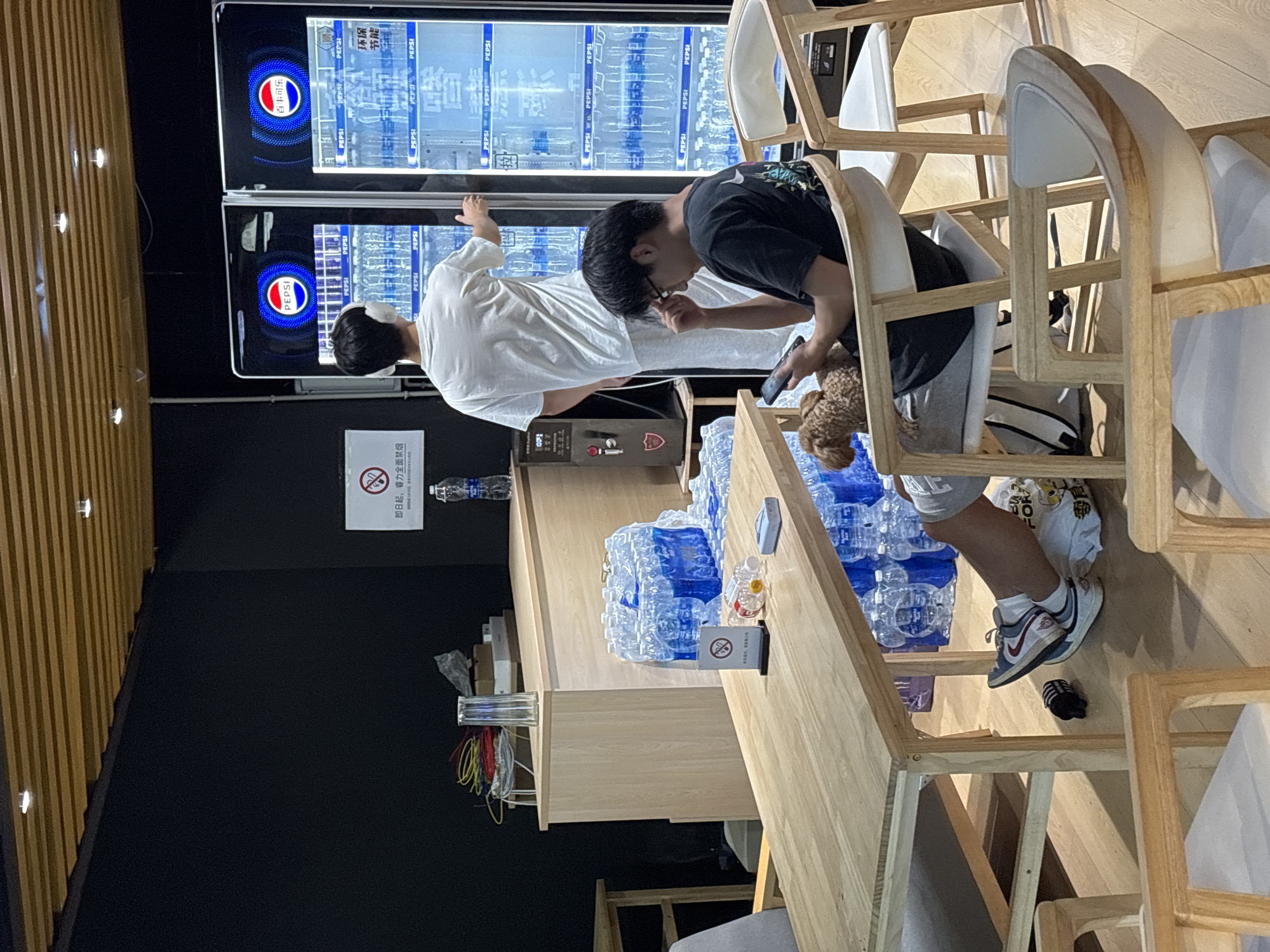 Here is a description of the image:
**Location:** The image appears to be taken indoors at a business or restaurant in the Nan'an District, China.
**Scene:** The scene is a casual setting, likely a break room or refreshment area.
**People:** There are two people in the image, both young men of East Asian descent.
* One is wearing a white shirt and stands at a Pepsi vending machine.
* The other is sitting in a wooden chair, wearing a black shirt and gray shorts. He is using a smartphone.
**Objects:**
* Pepsi vending machines: There are two large Pepsi vending machines stocked with bottled water.
* Wooden chairs and tables: There are several wooden chairs and a long wooden table.
* Bottled water: Numerous bottles of water are piled on the table.
* Smartphones
* Dog: A small, brown, poodle-type dog is seen by the chair.
**Actions:**
* One man is interacting with the Pepsi vending machine.
* The other man is sitting down and using his smartphone.
**Interactions:** The two men seem to be together, possibly taking a break. The second man has his attention on his phone while the first man interacts with the vending machine.
**Environment:** The setting is clean and well-lit. The walls are a combination of black and wood paneling.