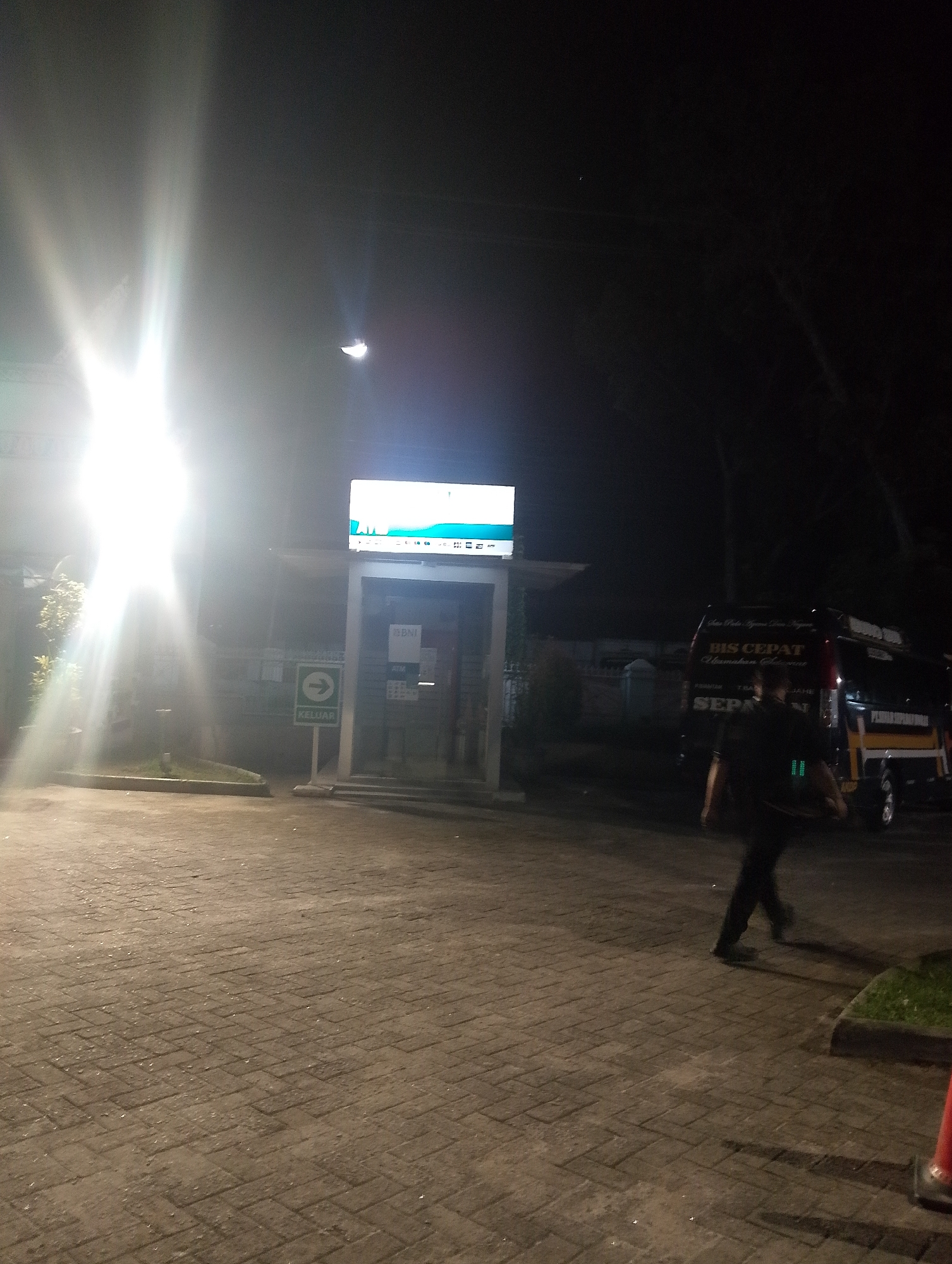 A nighttime exterior scene shows a paved walkway in front of a building. A bright light source emanates from the left of the frame, causing lens flare. A person walks across the walkway from right to left, silhouetted against the light. In the background, a dark-colored van with text on its side is parked. The building appears to be a bank, with signage indicating "BNI" and "ATM." A green directional sign with the word "KELUAR" (Indonesian for "Exit") and an arrow is also visible. The environment is dark with artificial lighting.