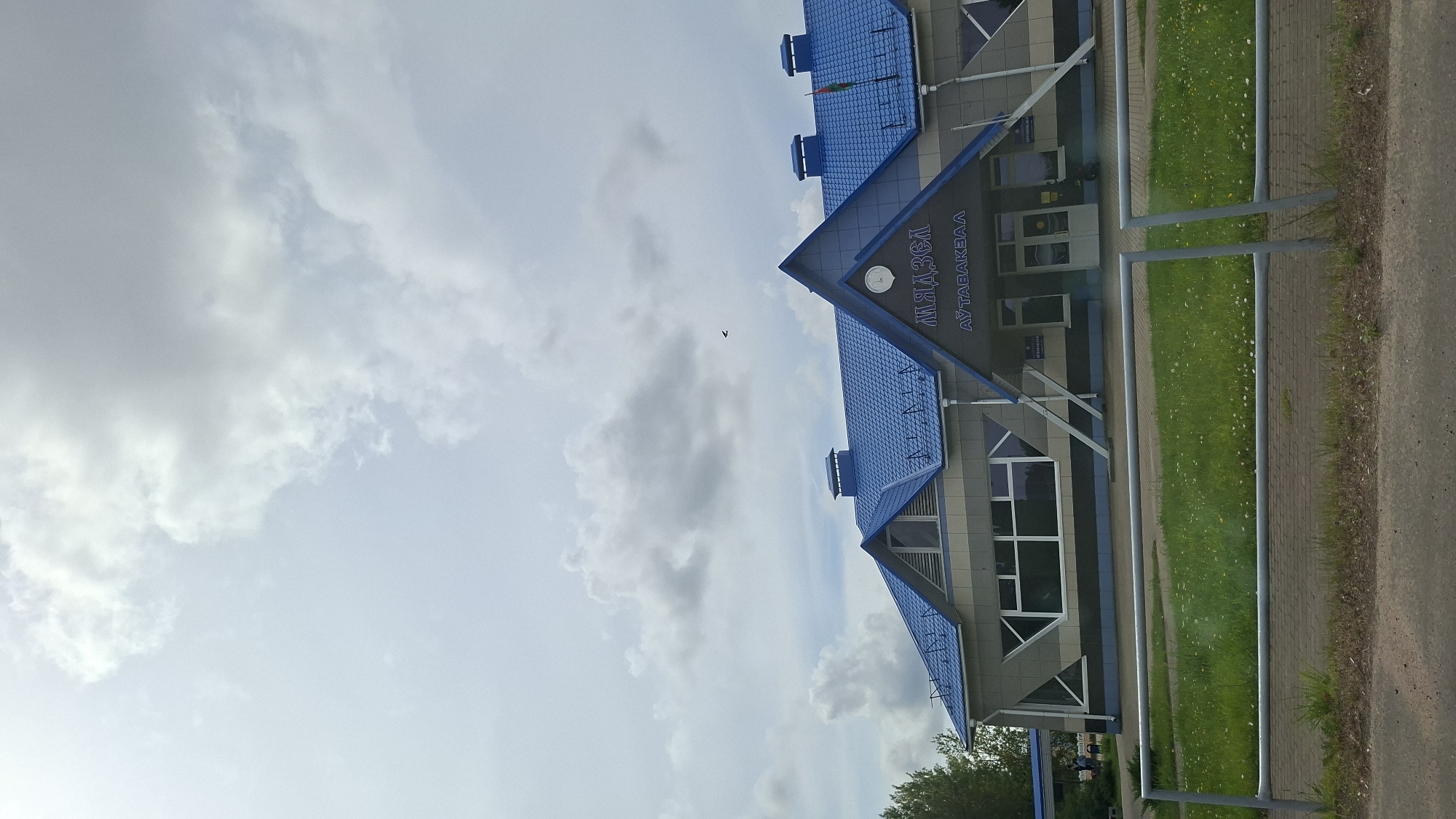 The image depicts the exterior of the Myadzyel bus station, identified by the Cyrillic lettering on the facade. The building features a modern design with a striking blue, multi-pitched roof. The facade is clad in a patterned material, primarily beige with grey and black accents. Large windows, some with geometric framing, are visible. Above the entrance, the words "МЯДЗЕЛ" (Myadzyel) and "АЎТАВАКЗАЛ" (Autavokzal, meaning bus station) are prominently displayed. A clock is mounted on the triangular section of the roof above the entrance. A Belarusian flag is affixed to the right side of the building. The bus station is situated in a green, grassy area with a metal fence in the foreground. The sky is partly cloudy with a bird visible in flight. The image appears to have been taken from a slightly elevated position, possibly from a vehicle.