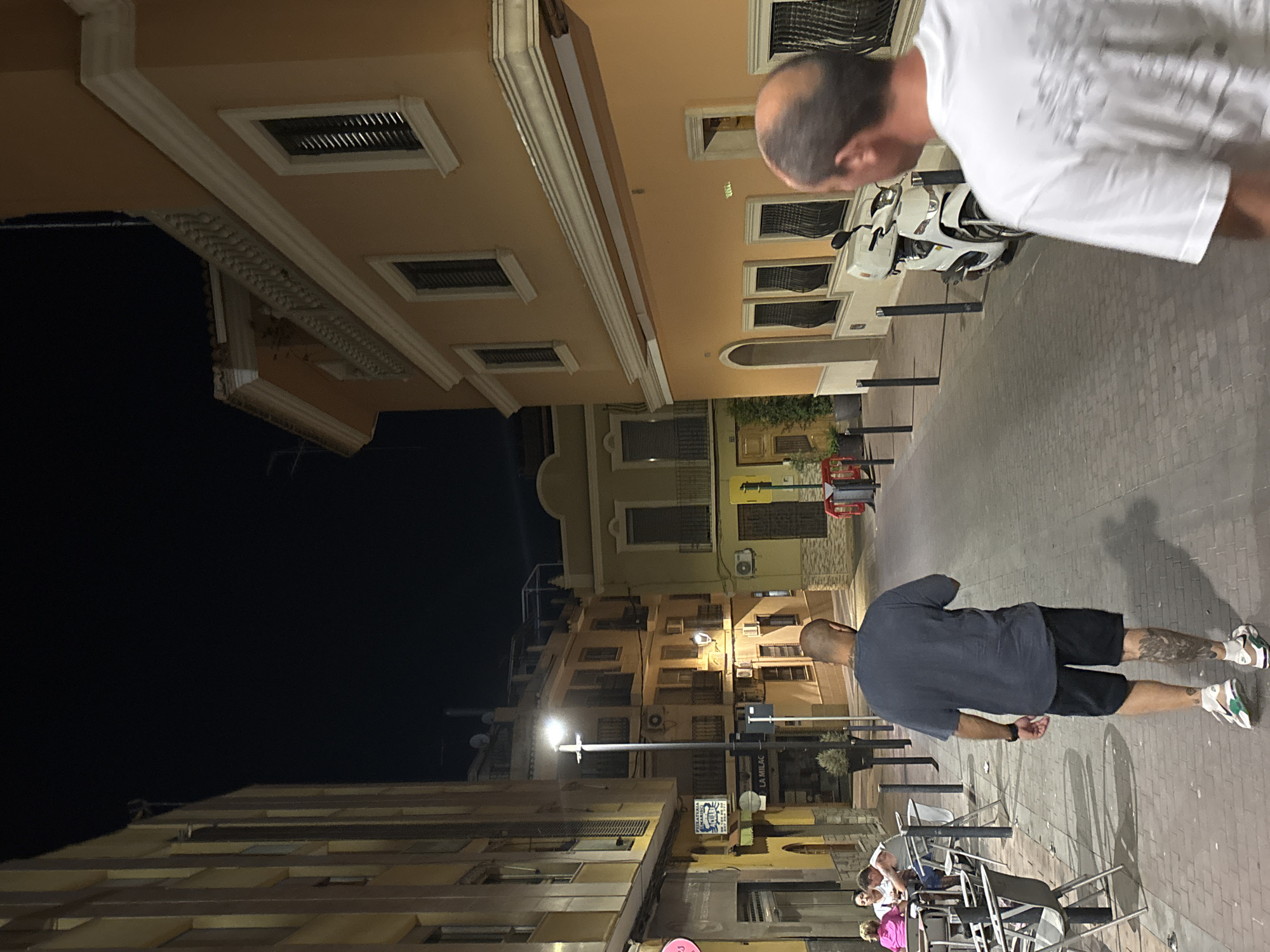 A low-angle, full-shot, nighttime street scene in Picassent, Spain, shows two men walking away from the camera on a brick-paved sidewalk. The man in the foreground is closer to the camera and wears a white t-shirt with a graphic print and a partially visible bald head. The man further down the sidewalk is in focus, wearing a dark blue t-shirt, black shorts, and athletic shoes, with visible tattoos on his legs.
The street is lined with buildings. On the left, a tall, cream-colored building with multiple windows and balconies is visible, partially obscured by shadows. On the right, an orange building with white trim and dark-shuttered windows dominates the view, featuring a balcony with ornate railing. A white scooter is parked along the sidewalk in front of the orange building.
Several outdoor tables and chairs from a restaurant or cafe are set up on the sidewalk on the left side of the street. In the distance, the street curves slightly, revealing more buildings and a streetlight. The overall atmosphere is that of a European town at night, with artificial lighting illuminating the street and facades.