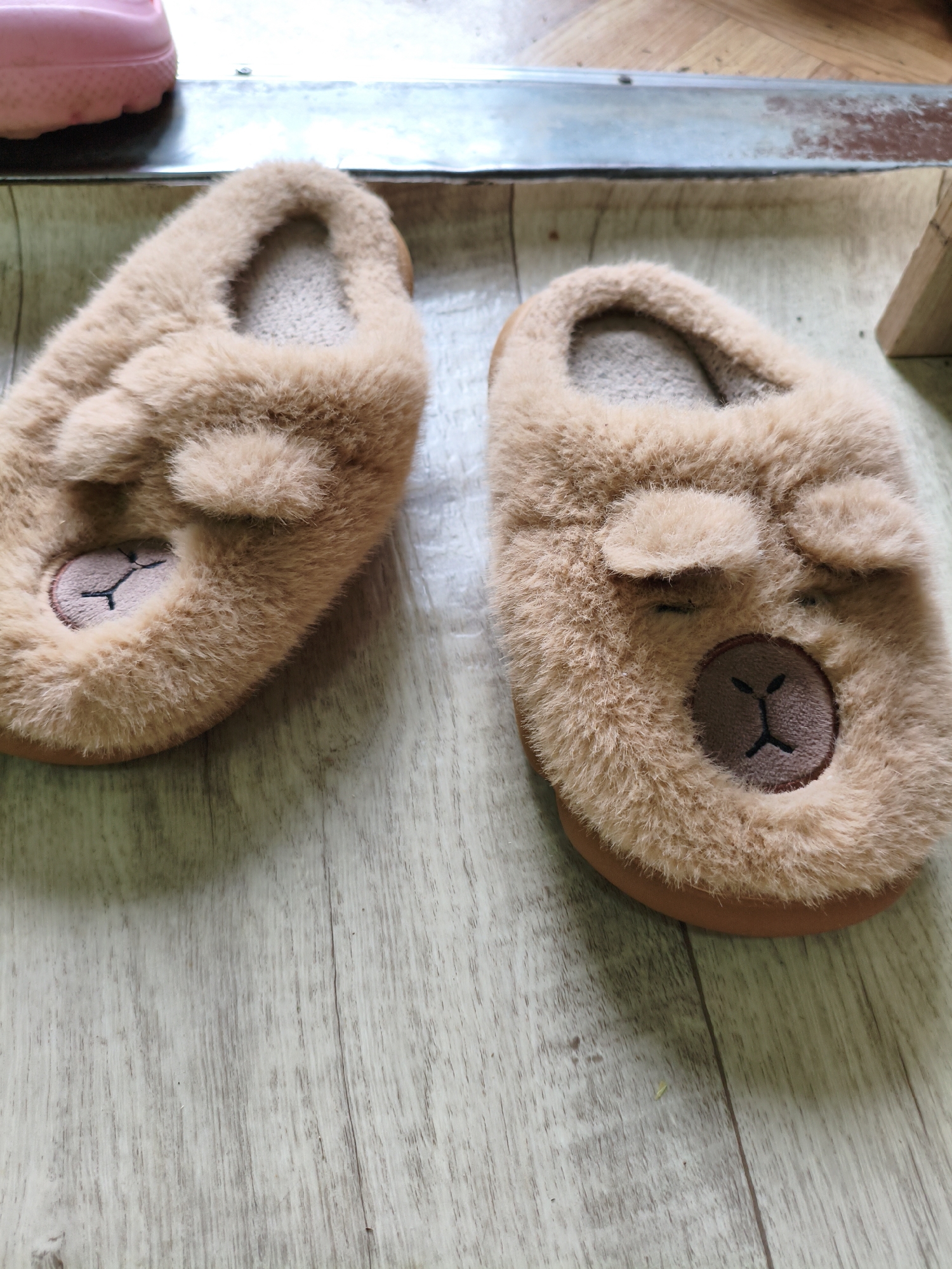 A pair of fuzzy brown bear-faced slippers rest on a light wood-grain floor. The slippers have small, round ears and embroidered facial features. A pink object, possibly another shoe or sandal, is visible in the upper left corner of the image. The slippers are not being worn. The scene is indoors, and no people are present. The image is shot from a slightly overhead angle.