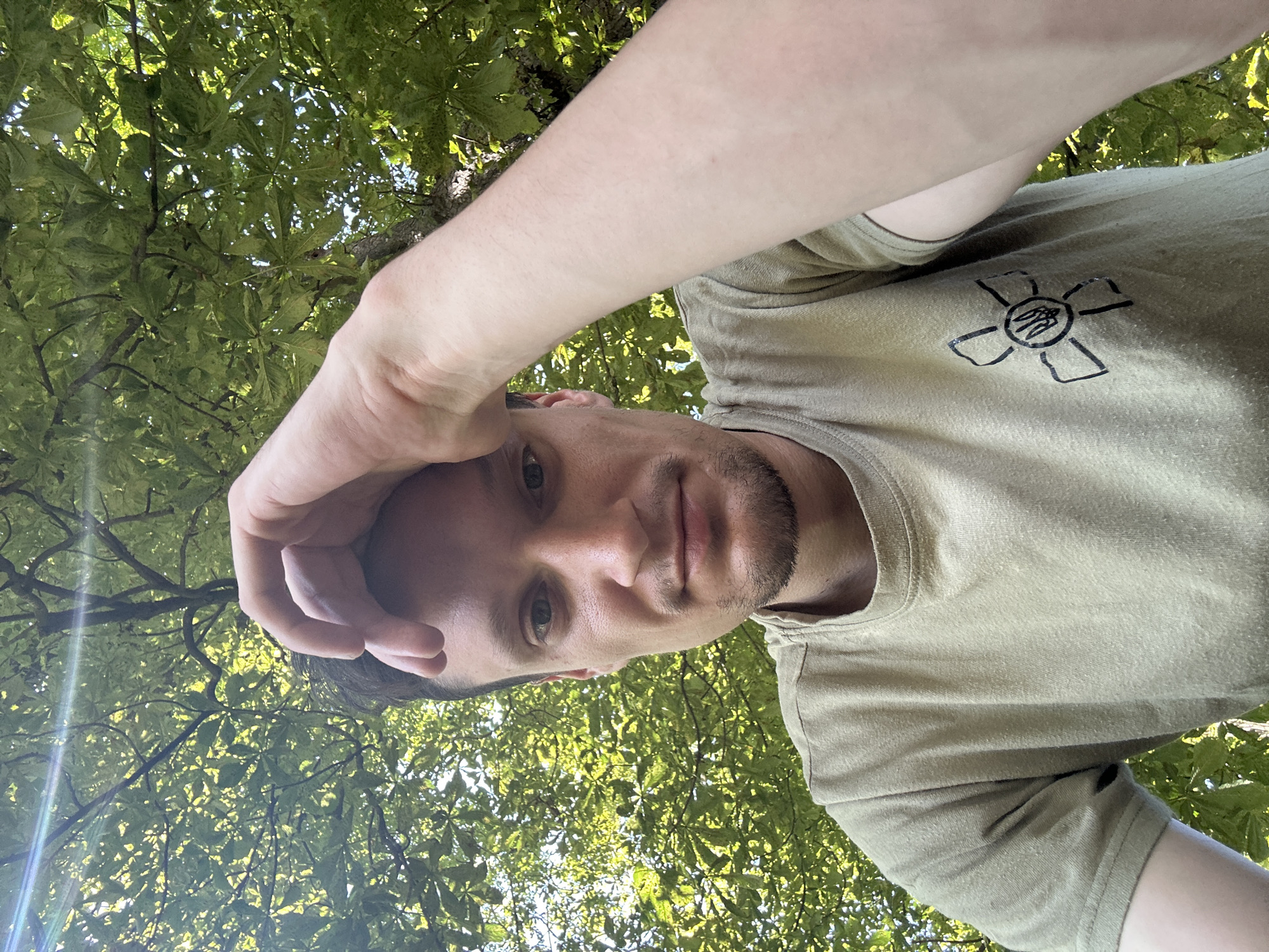 A medium-shot, outdoor selfie shows a man with short brown hair and light stubble. He is wearing a light olive-green t-shirt with a black emblem on the left breast, consisting of a stylized flower with a trident in the center. The man is looking directly at the camera, with his left hand resting on his forehead. Sunlight filters through the green leaves of trees overhead, creating a dappled effect and a lens flare in the upper left corner of the image. The background consists of dense foliage. The image appears to be taken in Makiv, Ukraine.