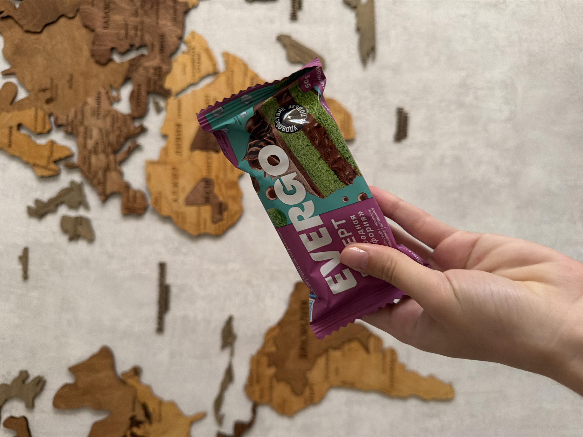 A hand, with light skin and a pale pink manicure, holds a teal and purple wrapper for a chocolate bar against a wooden map of the world. The wrapper features the brand "EVERGO" and an image of a slice of green cake with chocolate filling. Text on the wrapper is in Cyrillic, and some is blurred. The background is a wall with a wooden world map. The provided crops do not offer enough detail to determine the specific location within Semiluki, Russia.