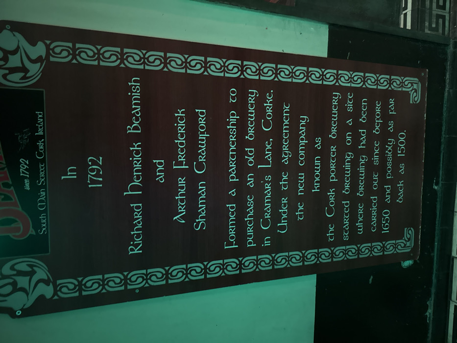 The image displays a vertically oriented dark brown wooden sign with light teal-green text and decorative Celtic knotwork borders. The sign is mounted on a light teal-green wall, above a dark horizontal surface. The top of the sign features text "since 1792" and "South Main Street Cork, Ireland." The main body of text describes a historical event: "In 1792 Richard Henrick Beamish and Arthur Frederick Shaman Crawford Formed a partnership to purchase an old brewery in Cramar's Lane, Corke. Under the agreement the new company known as the Cork Porter Brewery started brewing on a site where brewing had been carried out since before 1650 and possibly as far back as 1500." The scene is located indoors within a commercial establishment in the city of Paris, France.