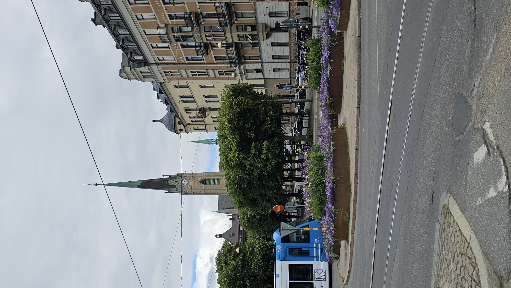 This is a street-level, medium-shot, eye-level, outdoor photograph taken on a cloudy day. In the foreground, a light grey asphalt road with two parallel tram tracks is visible, leading away from the viewer. A cobblestone sidewalk borders the road on the left.
Dominating the midground is a circular planter filled with purple and white flowers, with some vines cascading over the edge. Behind the planter, a blue and white tram is partially visible on the left. Further back, a line of lush green trees creates a natural barrier between the street and the buildings beyond.
In the background, a tall, slender church spire with a green roof and a clock face rises above the trees. To the right of the church, several multi-story apartment buildings with light yellow and cream-colored facades are visible. These buildings feature dark roofs, balconies with black railings, and numerous windows. A smaller green spire is also visible between the church and the apartment buildings.
The scene is set in a European city, likely Stockholm, Sweden, judging by the architecture and the presence of a tram. The environment appears to be a busy urban area with a blend of historic and modern buildings, public transportation, and green spaces. No people are clearly identifiable in the frame, except for a few indistinct figures in the distance. The overall impression is that of a typical city street on a day with overcast skies.