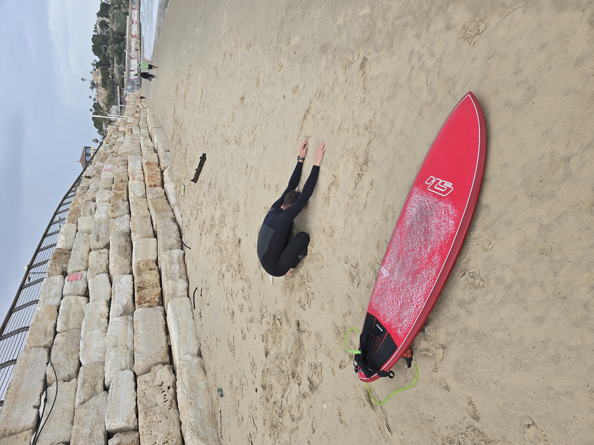 A man in a wetsuit stretches on the beach next to a red surfboard. The beach is sandy with footprints, and a retaining wall of large stones is visible on the left side of the image. A metal railing runs along the top of the wall. In the background, there is a glimpse of buildings and vegetation. The location is the beach in Tel Aviv, Israel.