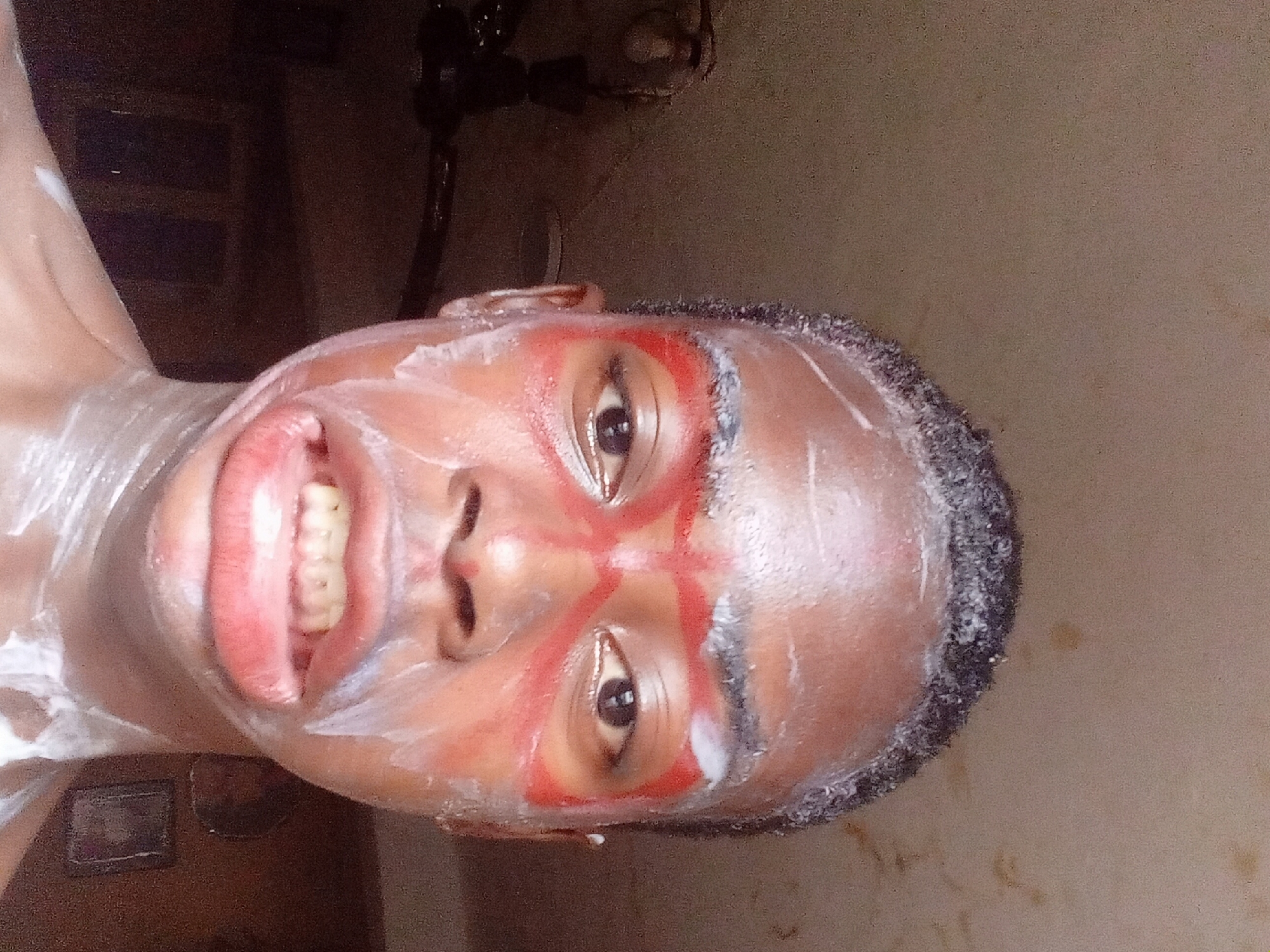 A close-up, portrait-oriented image shows a dark-skinned person with a shaved head, facing the camera with a wide smile, revealing teeth. The person's face is decorated with red markings around the eyes and a white substance covering parts of the face and upper body. The background is blurred, but a lamp or lighting fixture is visible. The setting is likely indoors, possibly a bathroom or similar space, based on the wall color and the presence of fixtures. The image was taken in Onitsha, Nigeria.
