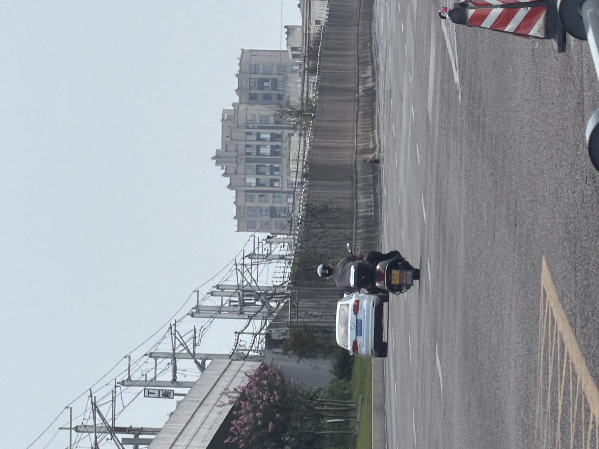 In the city of Jiangjin, China, on an overcast day, a motorcycle rider wearing a dark shirt and a helmet is moving away from the camera on a paved road. The rider is on a scooter with a yellow license plate. To the left of the motorcycle, a white sedan is parked, facing the same direction as the motorcycle. A concrete overpass with power lines runs along the left side of the image, with a sign displaying Chinese characters and a "T" symbol. In the background, a tall, weathered building with many windows stands behind a textured concrete barrier. The road is marked with white lines, and traffic cones are visible in the foreground on the right.