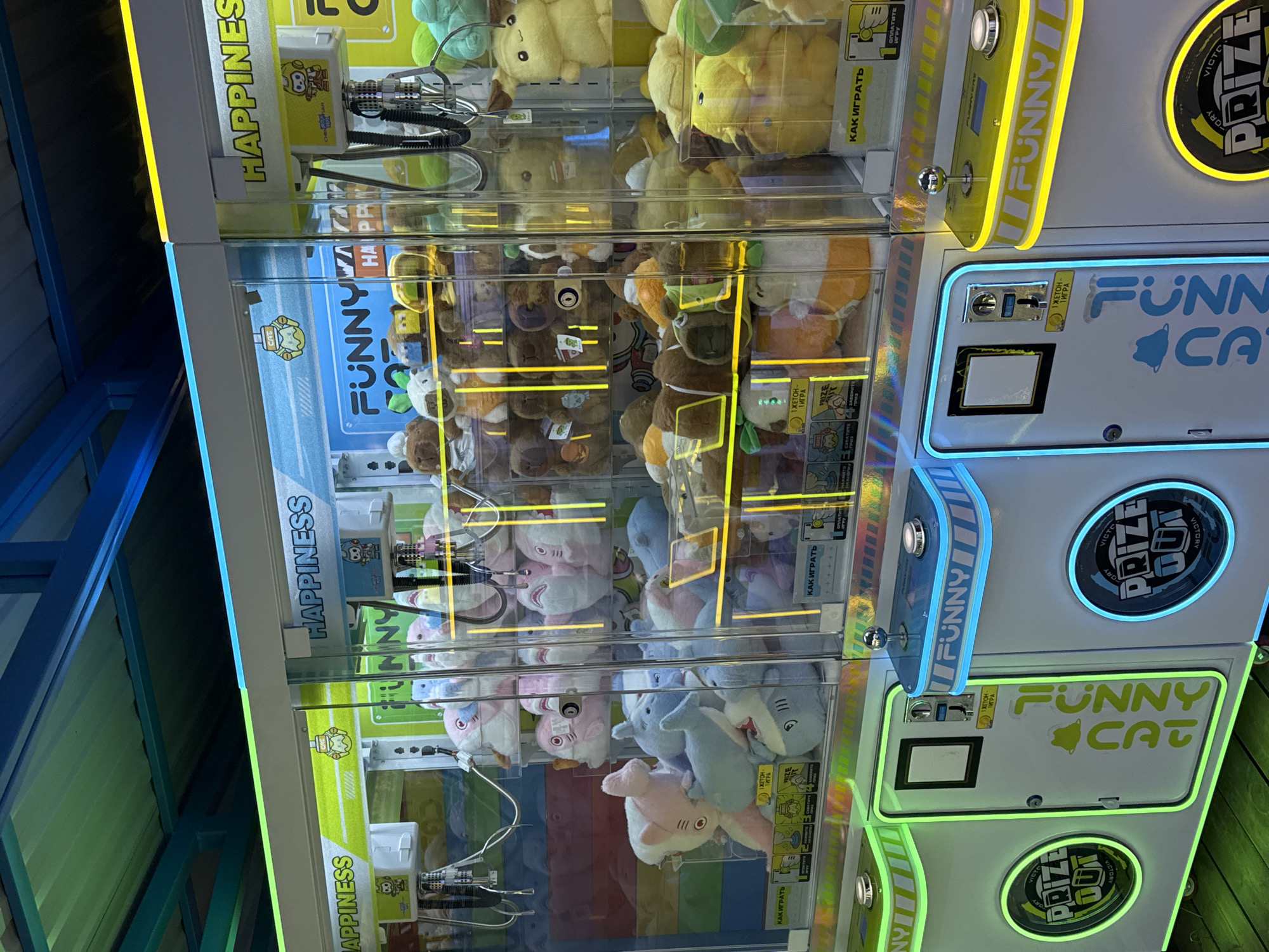 The image captures a vibrant, brightly lit indoor scene, likely an arcade or entertainment center in Chaikovsky, Russia, featuring multiple claw crane game machines. The machines are stacked vertically, showcasing clear glass fronts filled with a variety of plush toys. The perspective is slightly tilted, suggesting a handheld photograph.
The machines are adorned with eye-catching neon-style lighting in yellow, blue, and green, illuminating the control panels and prize collection areas. No people are visible, but the setup implies an environment for playing these skill-based games to win prizes.
Visible through the glass, the prizes consist of a diverse assortment of stuffed animals: numerous yellow plushies, various shades of brown teddy bears, and a collection of pink and blue plush sharks or marine creatures. The metallic claw mechanisms are clearly visible inside the machines, positioned above the prizes.
Several labels and instructions are present in both English and Cyrillic script. Recurring English text includes "HAPPINESS" on the upper sections of the machines and "FUNNY CAT" with a bell icon on the side panels. Circular illuminated signs prominently display "PRIZE OUT," often accompanied by "VICTORY" or "VICTOR." Cyrillic text provides operational guidance such as "КАК ИГРАТЬ" (HOW TO PLAY), "1 ЖЕТОН- ИГРА" (1 TOKEN - GAME), and "ОПЛАТИТЕ ИГРУ" (PAY FOR THE GAME). Reflections on the glass surfaces create a dynamic visual effect, mirroring parts of the machines and the surrounding environment.