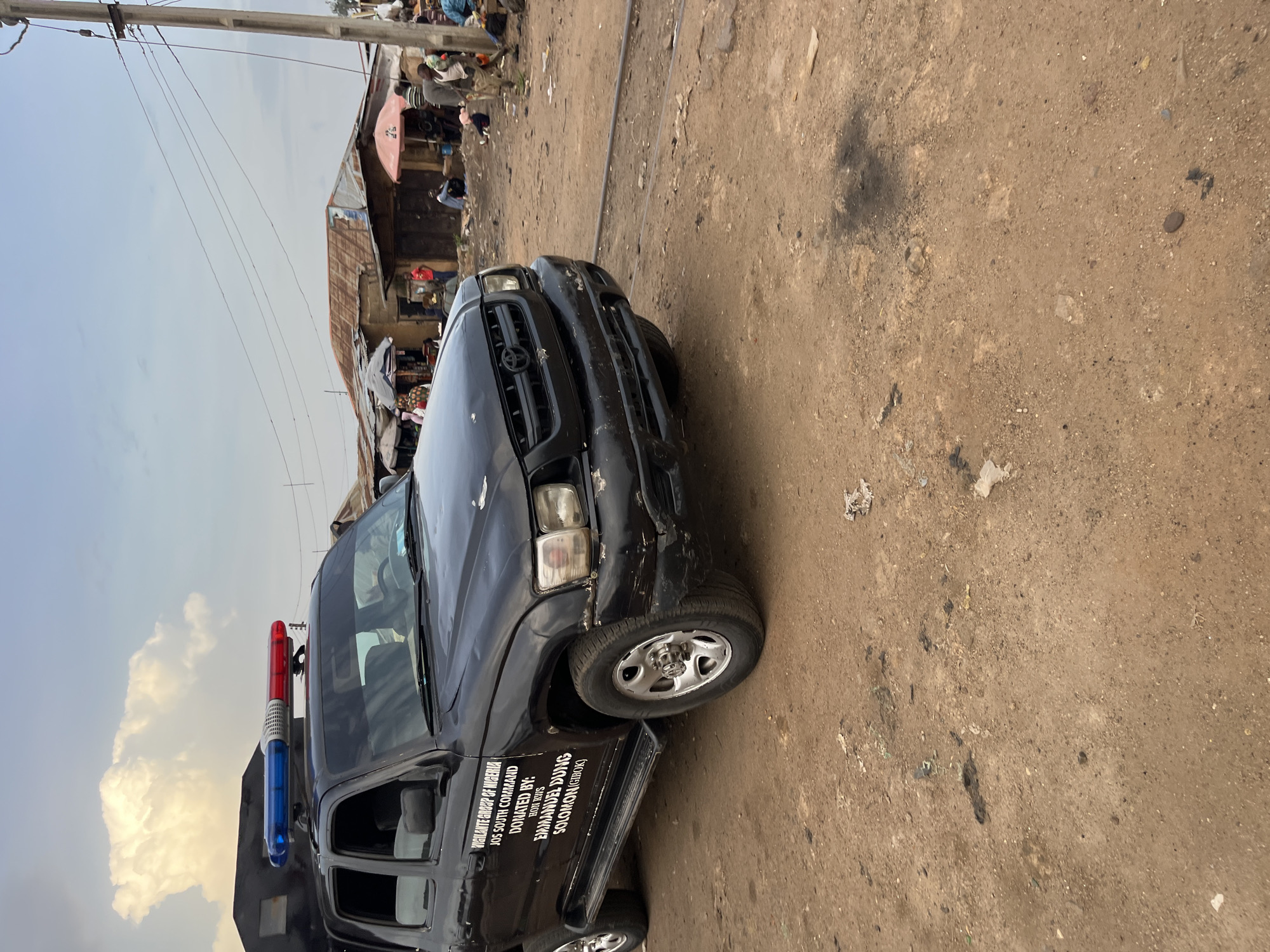 A black SUV, identified as belonging to the "Vigilante Group of Nigeria, Jos South Command," is parked on a dirt road in Bukuru, Nigeria. The vehicle has a red and blue light bar on its roof. The side of the SUV displays text identifying it as donated by "HON. KVIS EMMANUEL DUNG SOLOMON (GIROK)". In the background, a settlement with corrugated metal roofs is visible, along with several individuals gathered around market stalls. The environment is characterized by dusty terrain and overhead power lines.