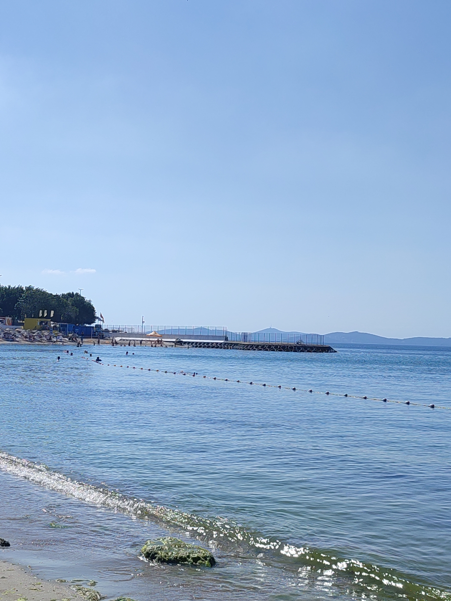 The image depicts a coastal scene, likely in Kadıköy, Turkey, with a clear blue sky overhead. In the foreground, gentle waves lap onto a sandy shore dotted with seaweed and a few rocks. A floating line of buoys demarcates an area of the sea, with several people swimming and wading within it. In the middle ground, a jetty or pier extends into the water, topped with a fence and what appears to be a bicycle. The background features a tree line with some structures and a distant hilly coastline. The overall atmosphere is tranquil and suggests a sunny day at the beach.