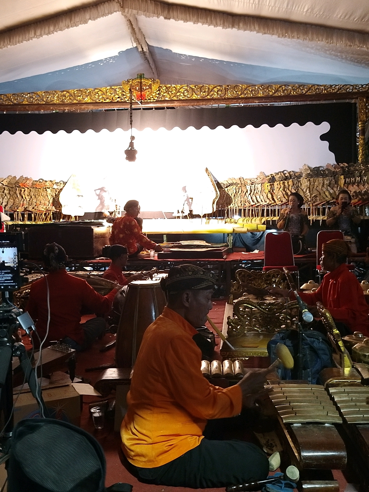 A group of musicians perform traditional Javanese gamelan music and wayang kulit shadow puppetry under a white tent in Putat, Indonesia. The scene is illuminated by stage lighting, casting shadows on the white screen behind the puppets. Several men are seated on the floor, playing various gamelan instruments, including xylophones and drums. One man in an orange shirt is actively playing a xylophone. In the background, three women are seated and appear to be singing or vocalizing. The stage is decorated with ornate golden carvings and a collection of wayang kulit puppets are visible. A camera on a tripod is positioned to the left, suggesting the performance is being recorded. The overall atmosphere is one of a cultural performance or festival.