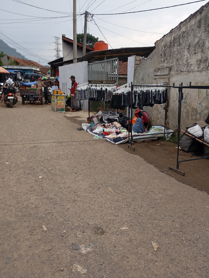 The scene is a street in Kutawaringin, Indonesia, located at latitude -6.974 and longitude 107.52. There are two people sitting on the ground, a man standing next to a rack of clothing, a motorcycle, a small cart, and a variety of other objects. The man is wearing a red shirt and jeans, and the two people sitting on the ground are wearing casual clothing. The ground is covered in loose gravel, and there is a small pile of clothing on the ground in front of the people sitting down. The clothing on the rack appears to be mostly jeans, and there are several other items, such as towels, bags, and shoes, piled on the ground next to the rack. There is a building in the background, and a large metal gate to the left of the clothing rack. The scene is likely a market or a small store.