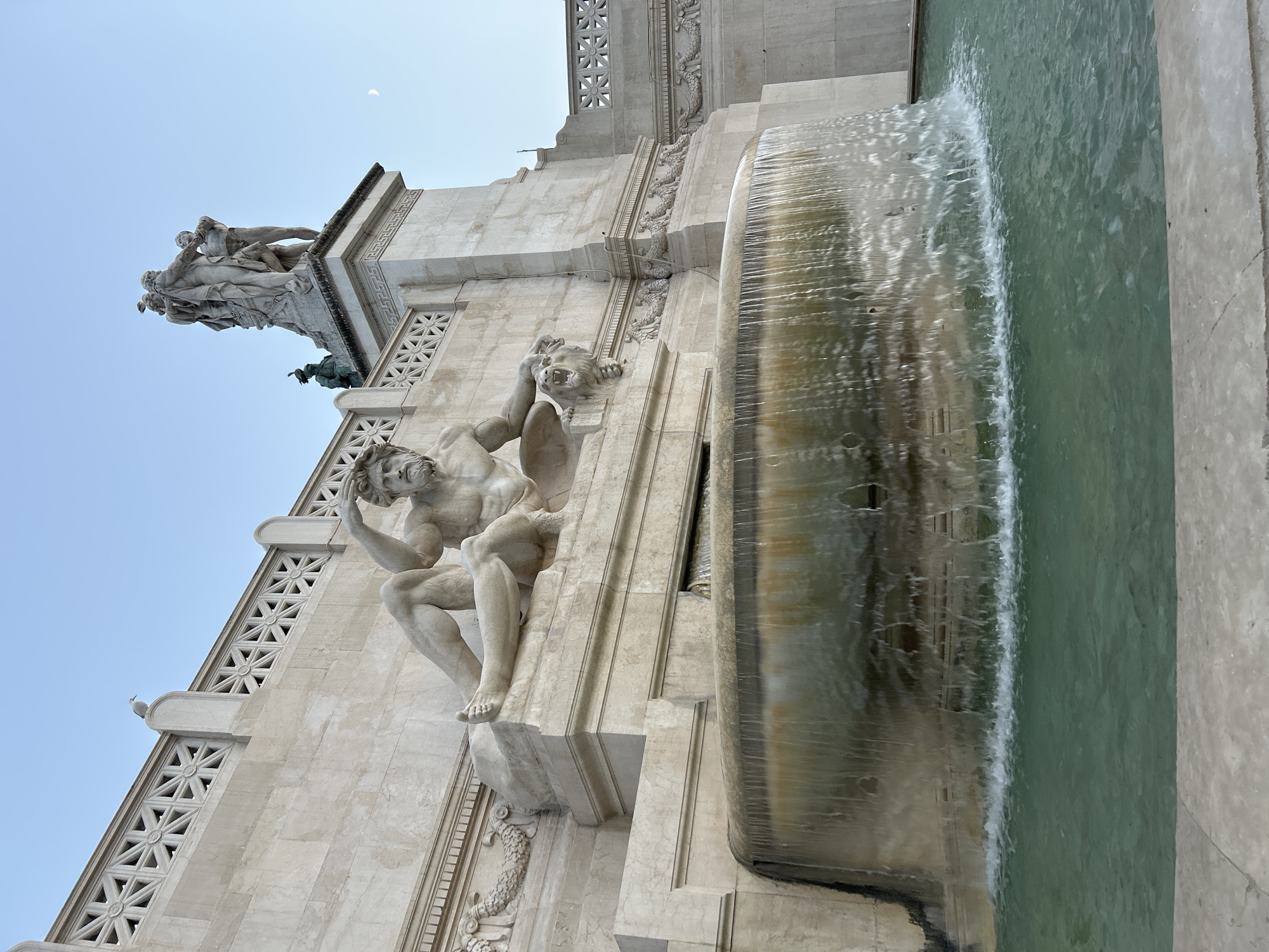The image shows the Fountain of the Sirens, part of the Altare della Patria (Altar of the Fatherland), also known as the Monument to Victor Emmanuel II, in Rome, Italy. The fountain features a large, circular basin with water cascading over its rim. To the left, a stone sculpture of a reclining male figure, possibly a river god or a mythological creature, is seated on a pedestal. The figure has a beard and is looking upwards. To the right of the fountain, a large, ornate marble structure with intricate carvings and architectural details rises. At the top of this structure, a group of marble statues depicts several figures. In the background, the sky is a clear, pale blue, with a faint crescent moon visible. A seagull is perched on the railing of the monument above the reclining statue.