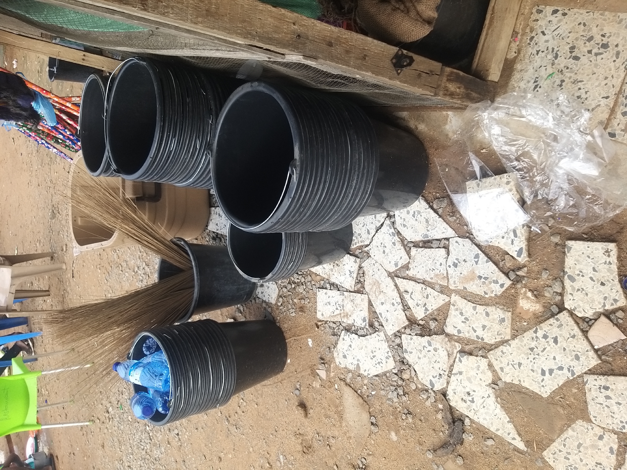 The image depicts an outdoor scene, possibly a marketplace or a storage area, with a focus on an assortment of black plastic buckets, brooms, and bottled water. In the foreground, several black plastic buckets are stacked, with one on the left containing two plastic bottles of blue liquid, likely water, and another bucket to its right is filled with dried broomsticks. Behind these, more stacked black buckets are visible. To the right of the buckets, a rough wooden structure with a wire mesh panel is present, adjacent to a large tan bucket or bin containing more broomsticks. Scattered on the ground are pieces of broken tiles, soil, and gravel. In the background, elements of a city environment are faintly visible, including furniture (a green plastic chair and a light-colored stool), and parts of people's legs and shoes, suggesting human presence. The overall impression is one of organized clutter, perhaps a vendor's stall or a storage area. While the exact location in Kano, Nigeria, cannot be definitively determined from the visual evidence alone, the style of the brooms and the general outdoor market aesthetic are consistent with scenes found in such urban centers.