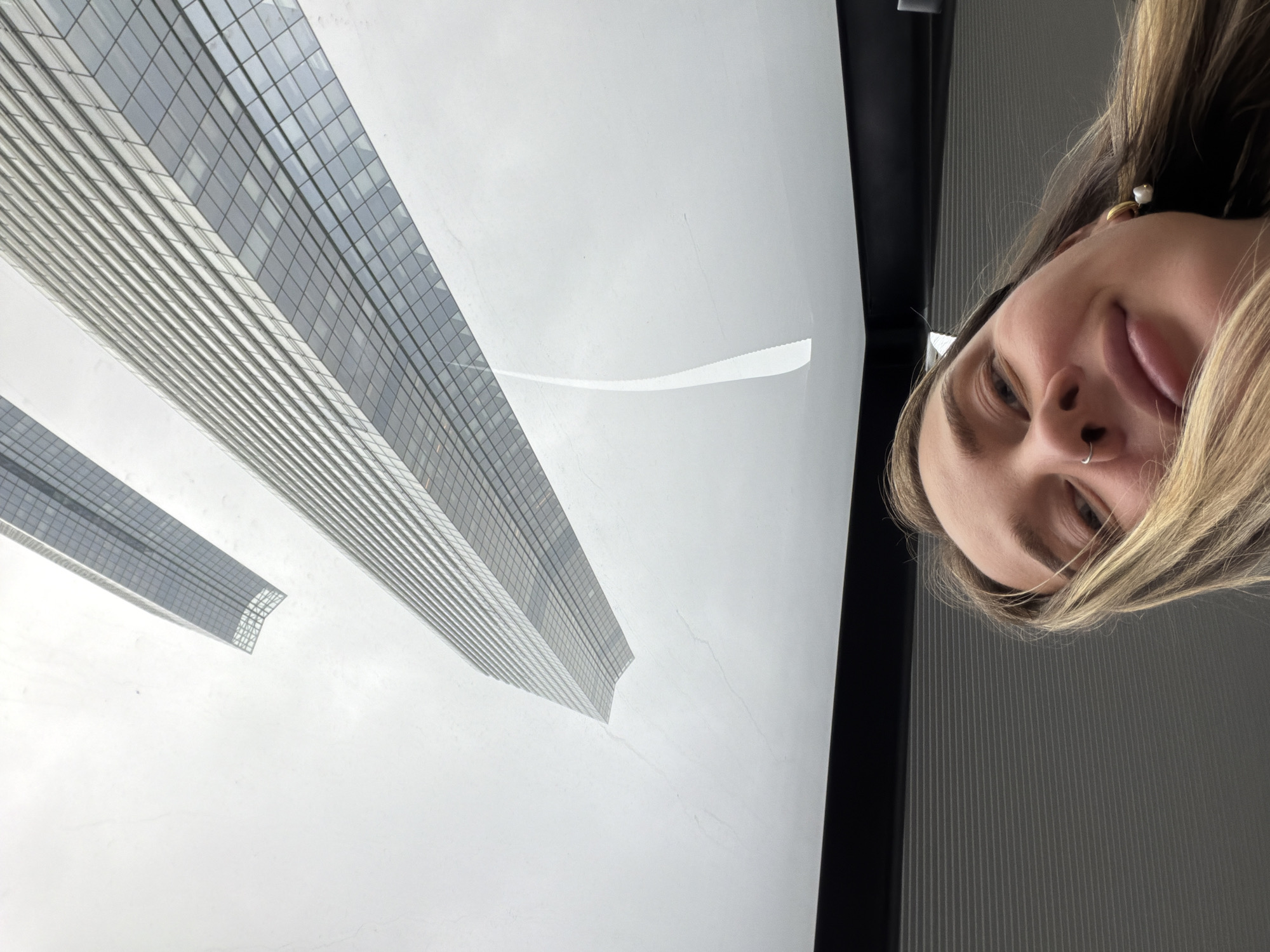 The image features a selfie of a woman with fair skin, blonde hair, and a nose piercing. She is wearing a pearl earring and her face is angled to the right side of the frame. In the background, visible through a window, are two skyscrapers. These are located in Moscow, Russia, specifically in the Moscow International Business Center, known as "Moscow City". The sky appears to be overcast or hazy, and the skyscrapers are depicted in tones of grey and white. The reflection of a portion of a wall or structure is also visible on the window.