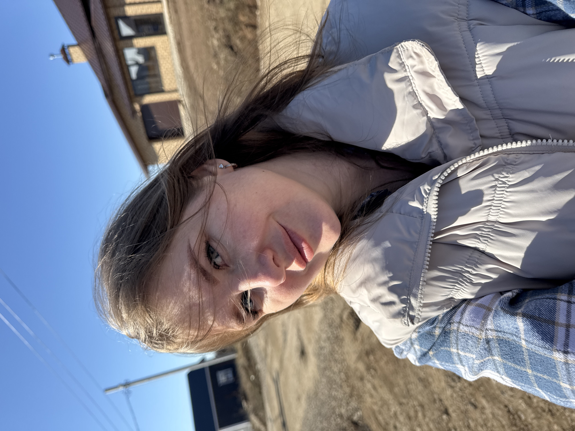 A young woman is posing outdoors in Gornyi (Dvurechenskoe S/P), Russia. She is wearing a light grey puffy jacket over a blue and white plaid shirt. The location appears to be near a building with brown brickwork under a clear blue sky with utility wires. The ground is light brown. The angle suggests the picture was taken from a low vantage point, looking slightly upwards.