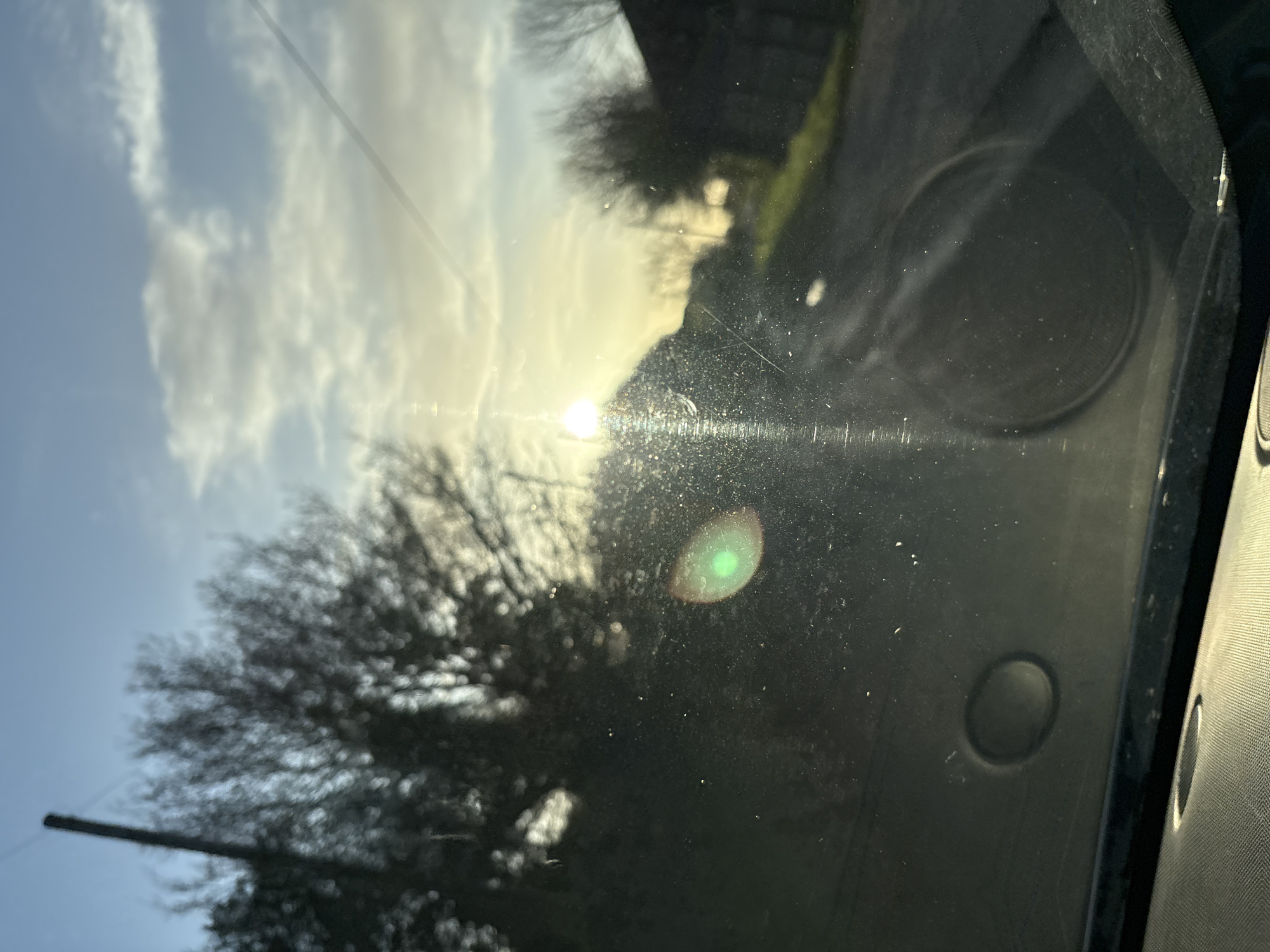 The image captures an exterior scene in Ruyton-XI-Towns, United Kingdom, viewed through the dirty and scratched windshield of a vehicle. The low sun, positioned in the upper right quadrant, causes significant lens flare, visible as a prominent green circular artifact in the lower-center and a vertical streak of light. The sky above is clear blue with scattered white clouds, transitioning to a bright golden hue near the horizon due to the sunlight.
In the immediate foreground, within the vehicle, the top edge of a textured dashboard is visible. A circular, black recessed object is present on the lower-left, while a circular speaker grille is visible through the windshield on the lower-right. The windshield itself exhibits numerous dust particles, smudges, and fine scratches.
Outside the vehicle, the environment is out of focus due to the focal point being on the windshield. On the far left, a dark, vertical pole is silhouetted against the sky, partially obscuring a large, bare-branched deciduous tree. A narrow, unpaved or gravel road or track extends into the background on the right, flanked by areas of green grass. Further into the background, indistinct, dark building structures and additional bare trees are silhouetted against the bright sky. No individuals, animals, or active subjects are visible in the scene.